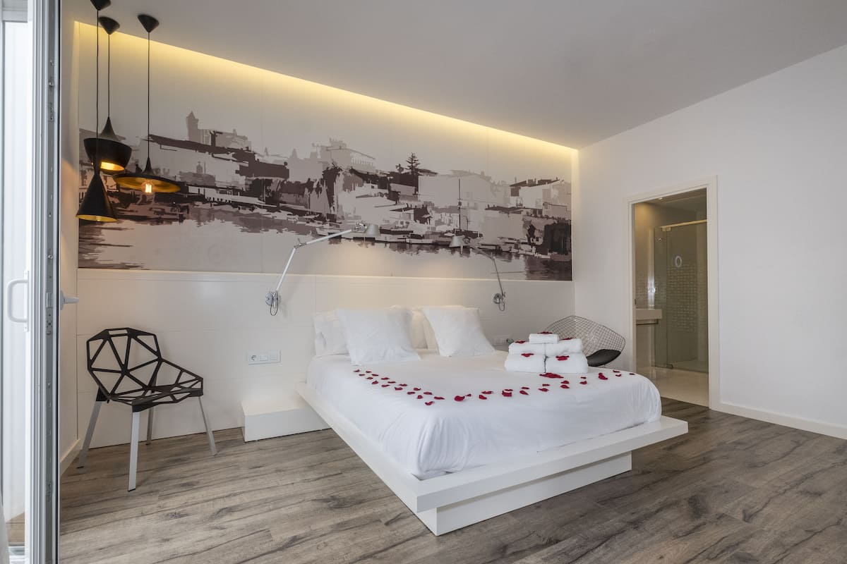 Un dormitorio moderno con una cama blanca, un mural de paisaje urbano, una silla geométrica y una puerta de ducha de cristal.