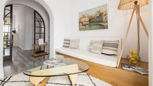 A modern living room with a glass coffee table, white sofa with patterned cushions, a painting of a coastal town, and a floor lamp with a woven shade. - Hotel Abril 37 (Ciutadella de Menorca)