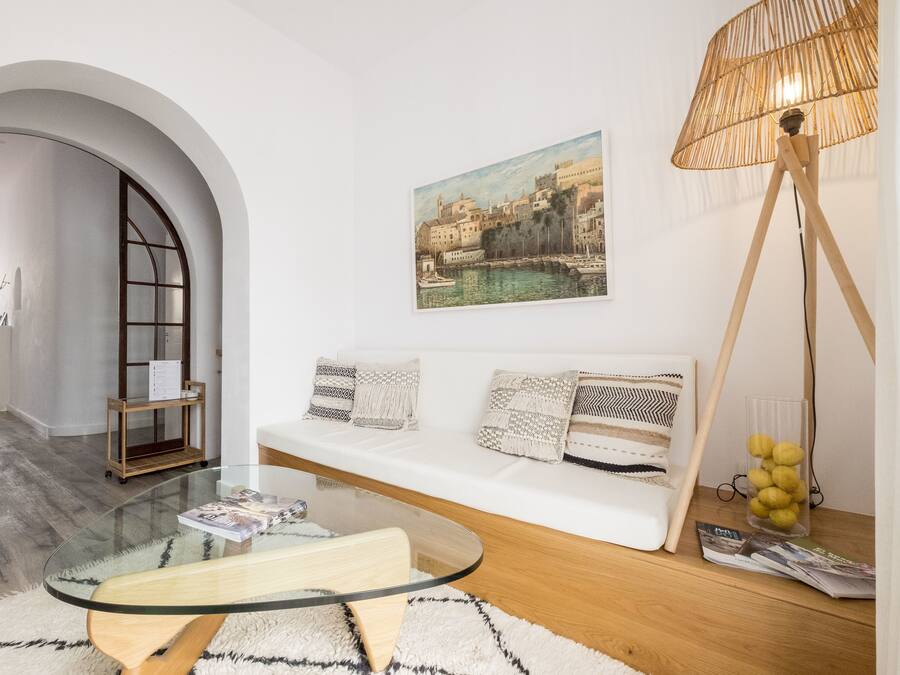 Un salon moderne avec une table basse en verre, un canapé blanc agrémenté de coussins à motifs, un tableau représentant une ville côtière et un lampadaire avec un abat-jour en tissu tressé.