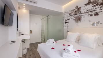 Ein Hotelzimmer mit einem Bett, weißen Handtüchern, einer Dusche, einem an der Wand befestigten Fernseher und einem Wandbild mit Stadtansicht.
