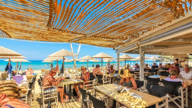 An outdoor dining area with tables and chairs under a thatched roof, overlooking a beach with umbrellas and people. - Venus Resort (Zakynthos)