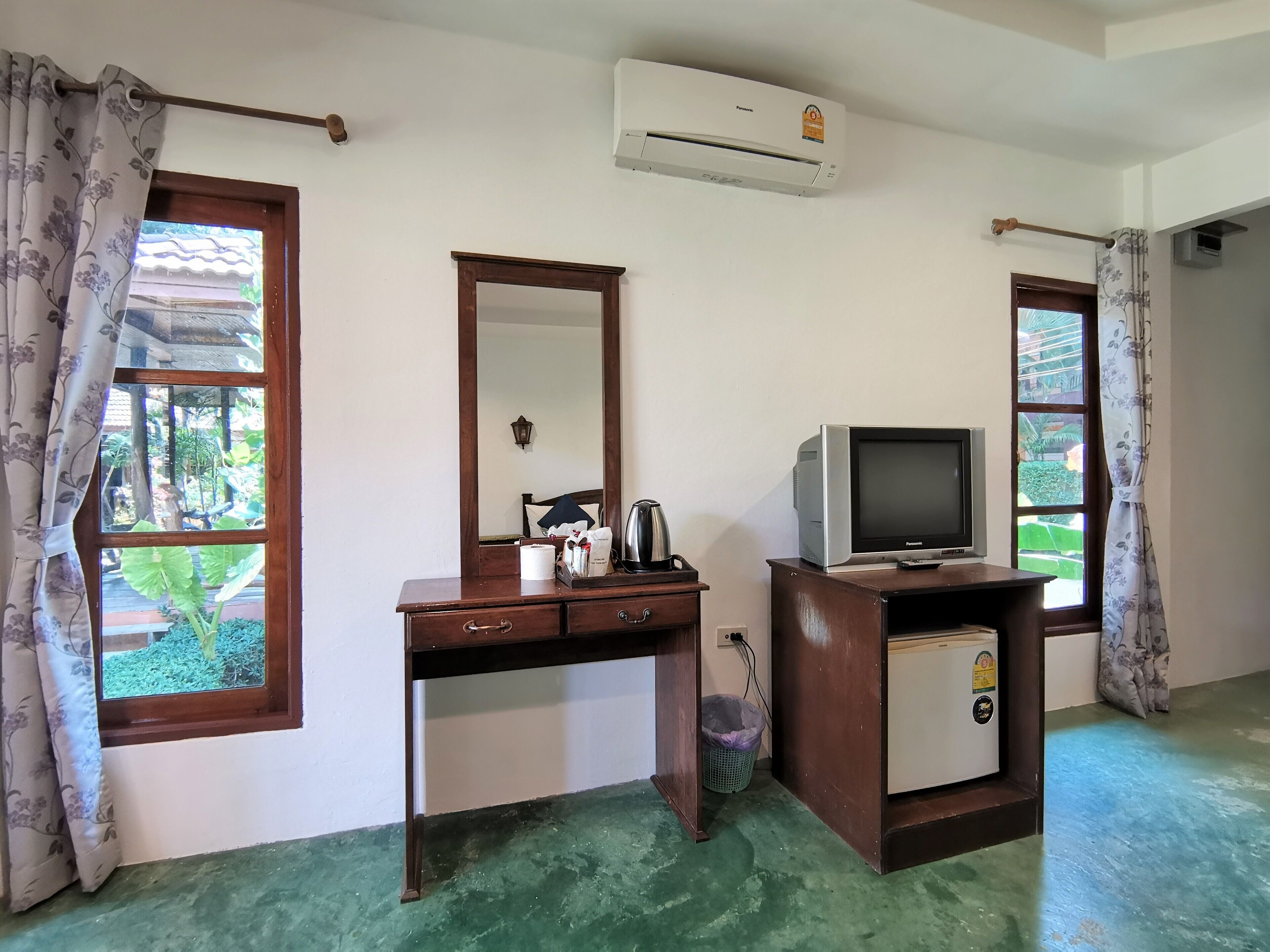 A room with a wooden desk, a mirror, a television on a stand, and an air conditioning unit.