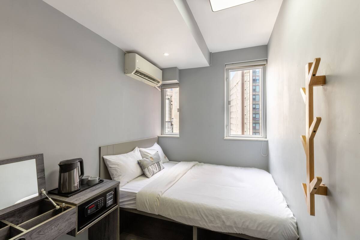 A modern hotel room with a bed, bedside table, air conditioning unit, and a window with a view of buildings.