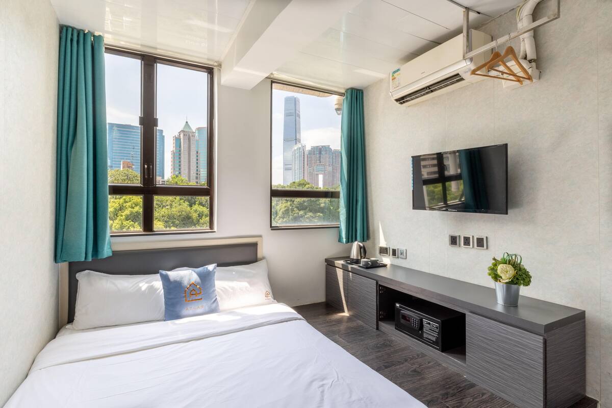 A modern hotel room with a large bed, a TV mounted on the wall, and a city view through the windows.
