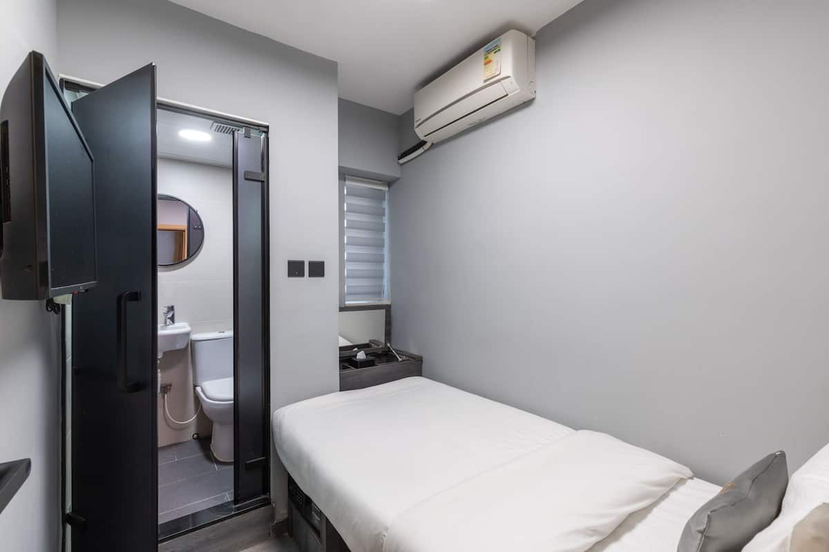 A compact room with a bed, a small bedside table, a wall-mounted air conditioner, a window with blinds, and a door leading to a bathroom.