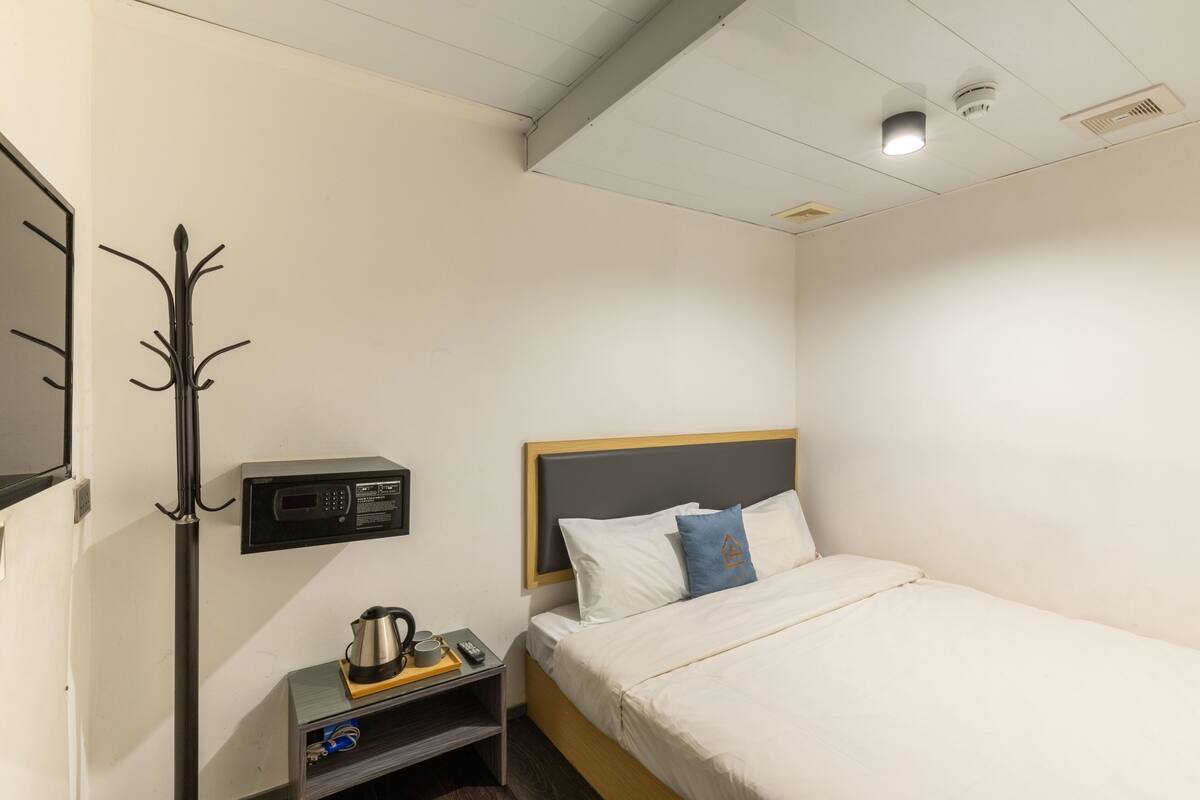 A hotel room with a single bed, a nightstand with a kettle and remote control, a coat rack, and a wall-mounted control panel.