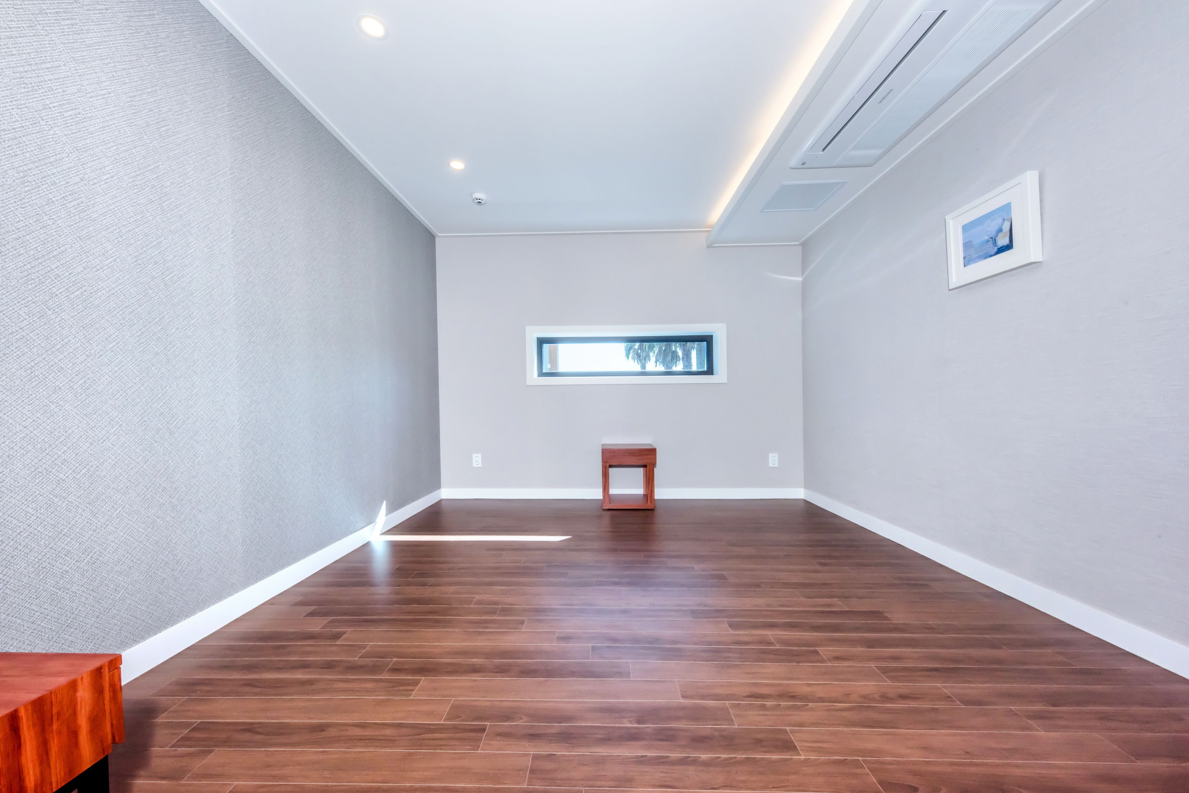 Une pièce spacieuse avec un plancher en bois, une petite table en bois, une fenêtre donnant sur un beau paysage et un tableau encadré accroché au mur.