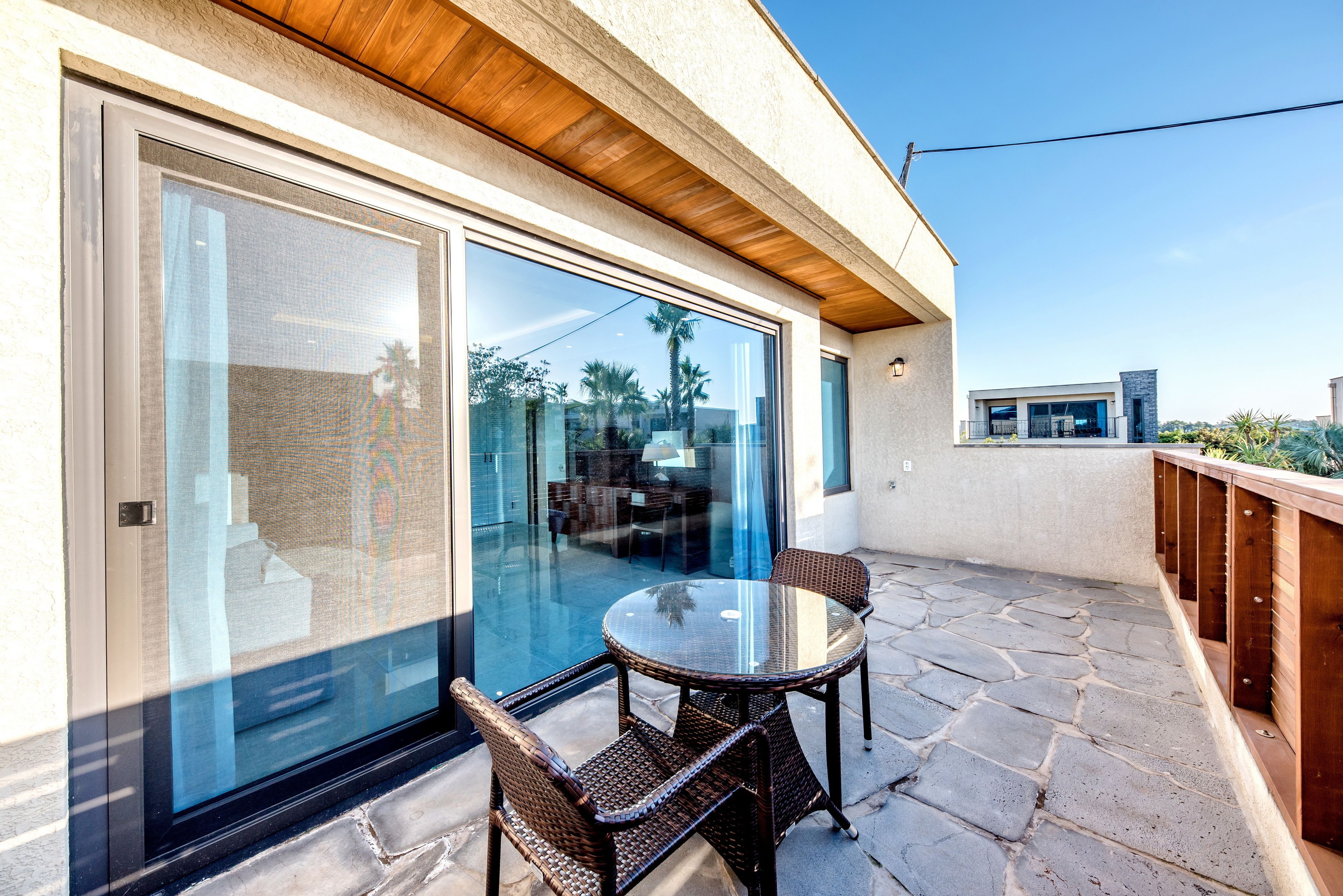 A glass-enclosed patio with a round table and wicker chairs, a stone-tiled floor, and a view of the surrounding landscape.
