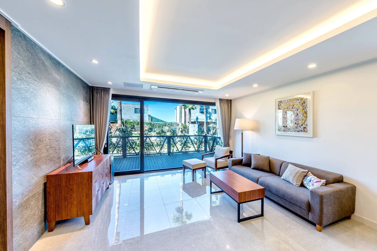 A modern living room with a sofa, coffee table, and TV stand. There is a balcony with a view of buildings and trees.