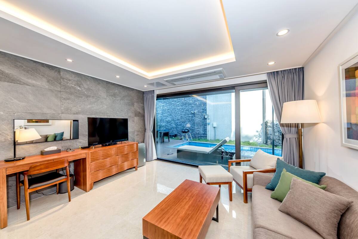 A modern living room with a wooden desk, sofa, and a TV. There is a large window with a view of a swimming pool and a stone wall.