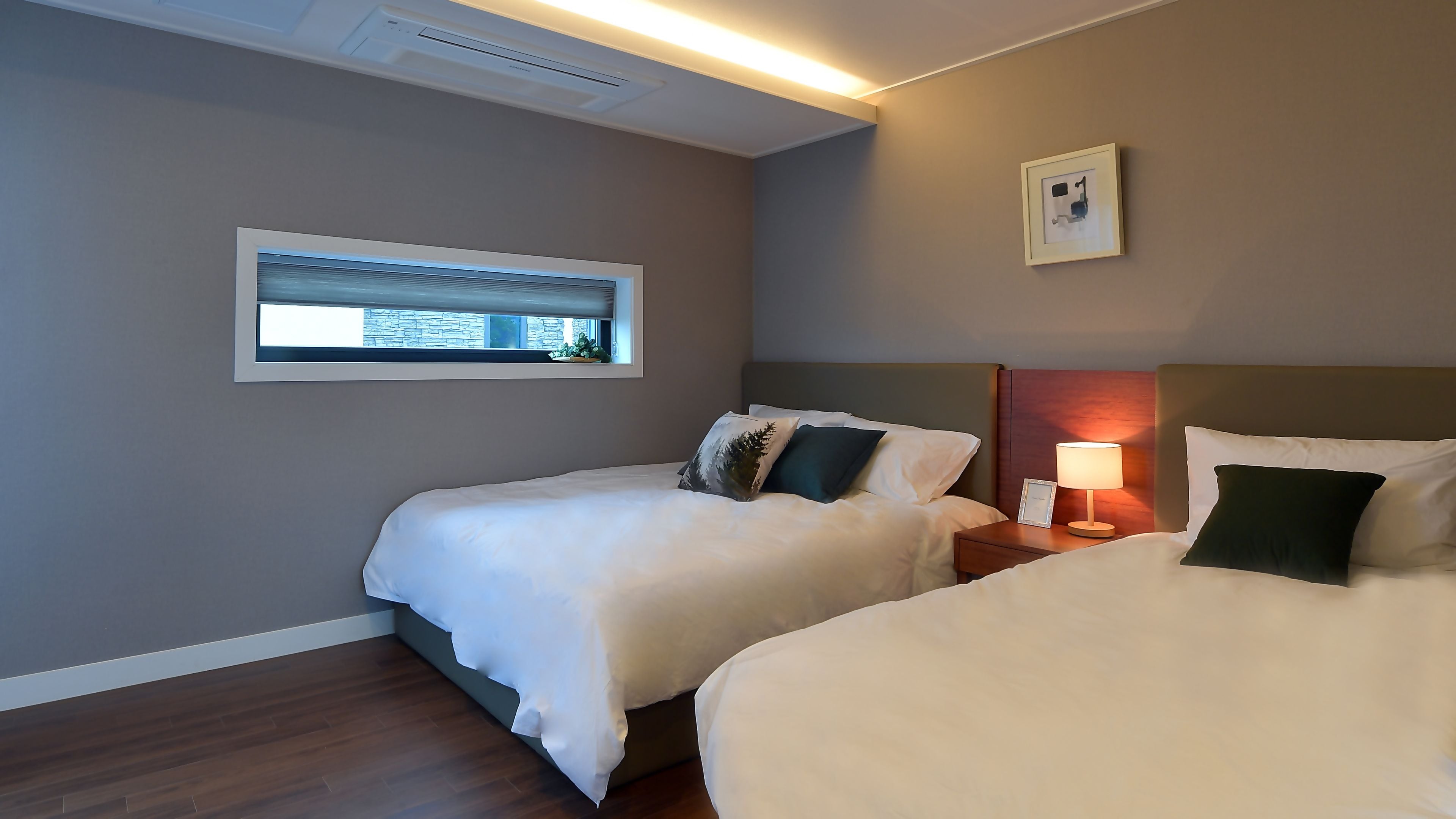 A hotel room with two beds, a bedside table with a lamp, a framed picture on the wall, and a window with a view of buildings.