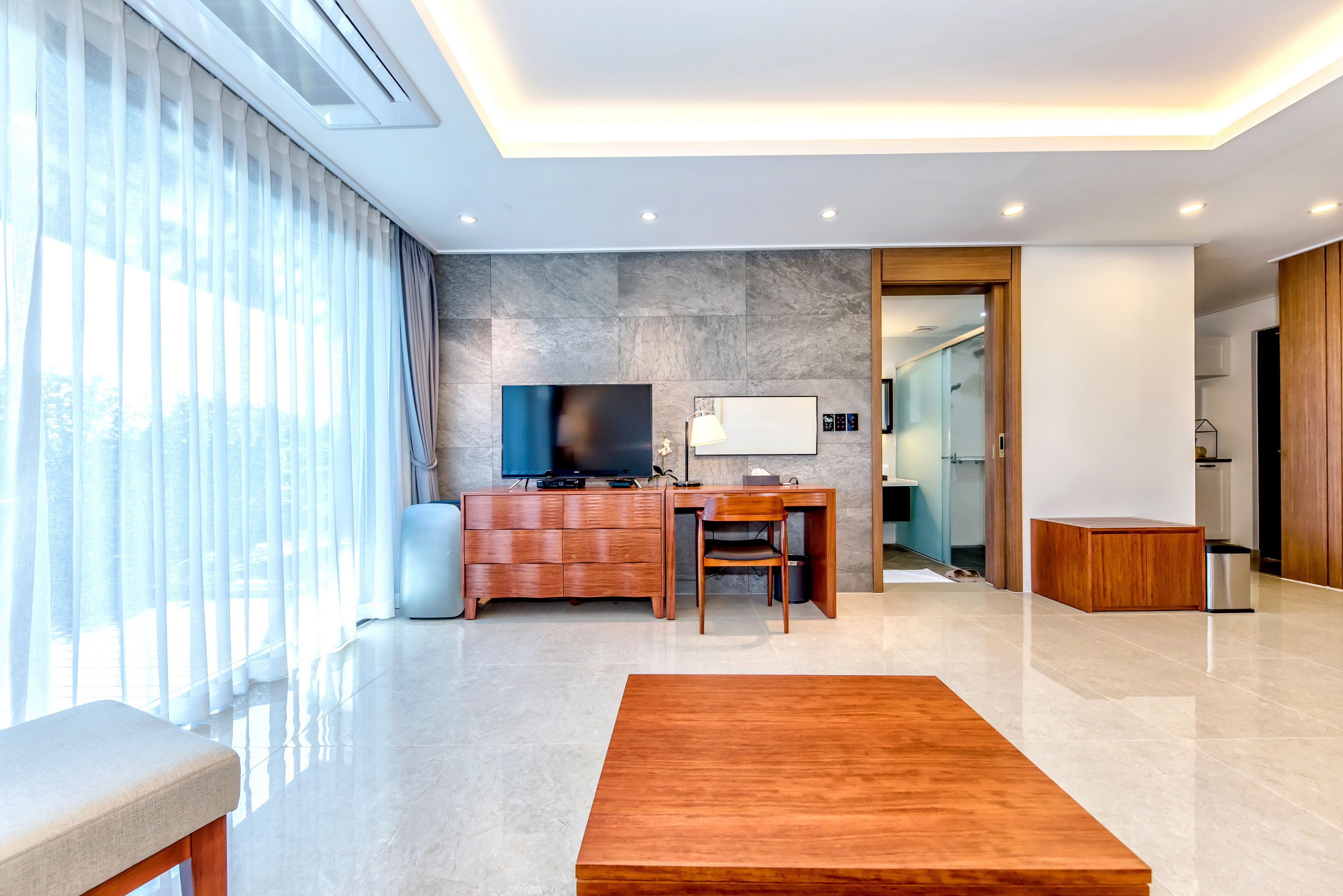 Una habitación de hotel moderna con un ventanal grande, un escritorio de madera, un televisor de pantalla plana y una zona de estar con un banco de madera.