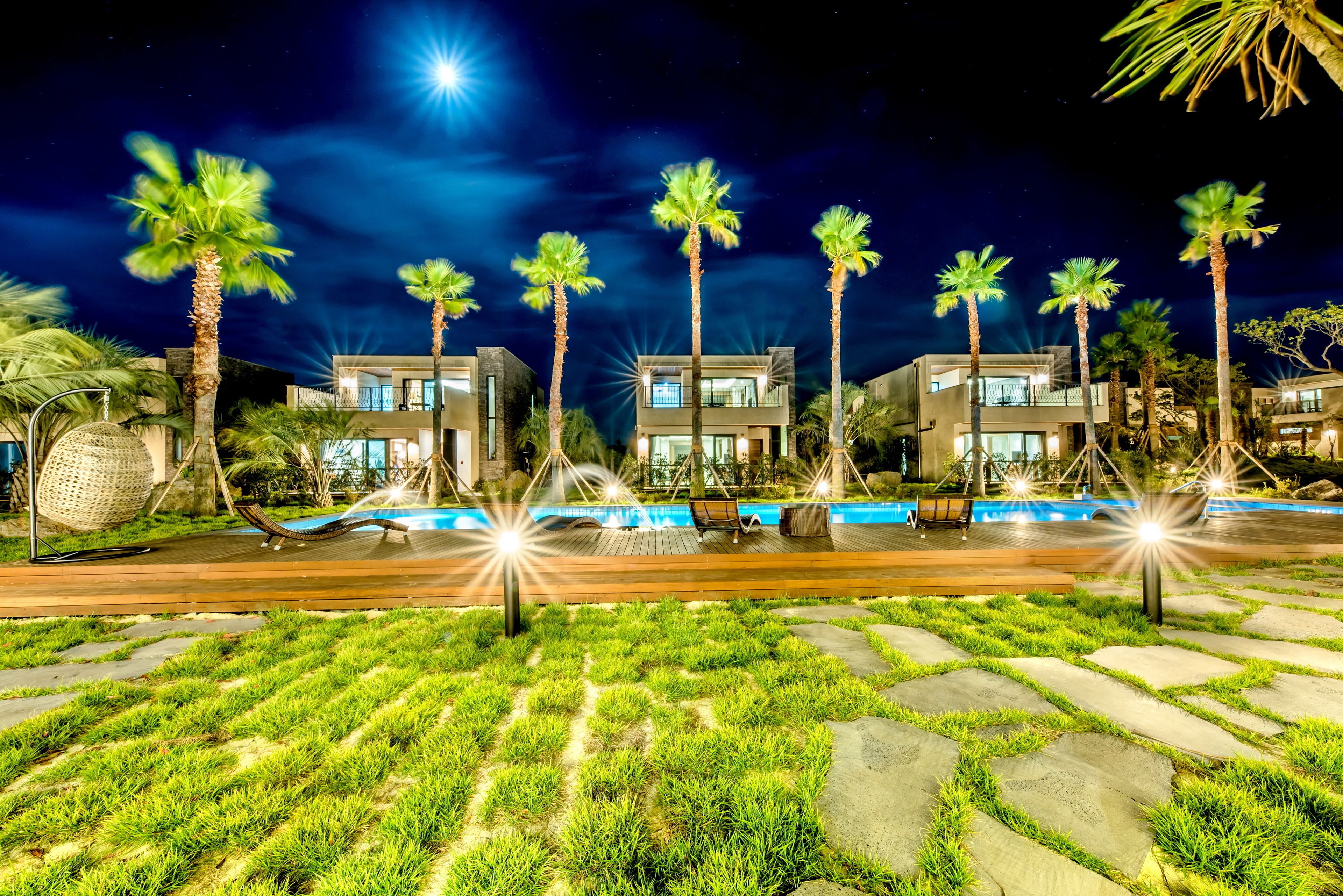 Un resort nocturno con edificios modernos, palmeras, una piscina y un paseo marítimo bien iluminado.