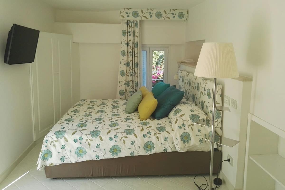 Un dormitorio con cama, televisor montado en la pared, lámpara y ventana con vista a follaje.