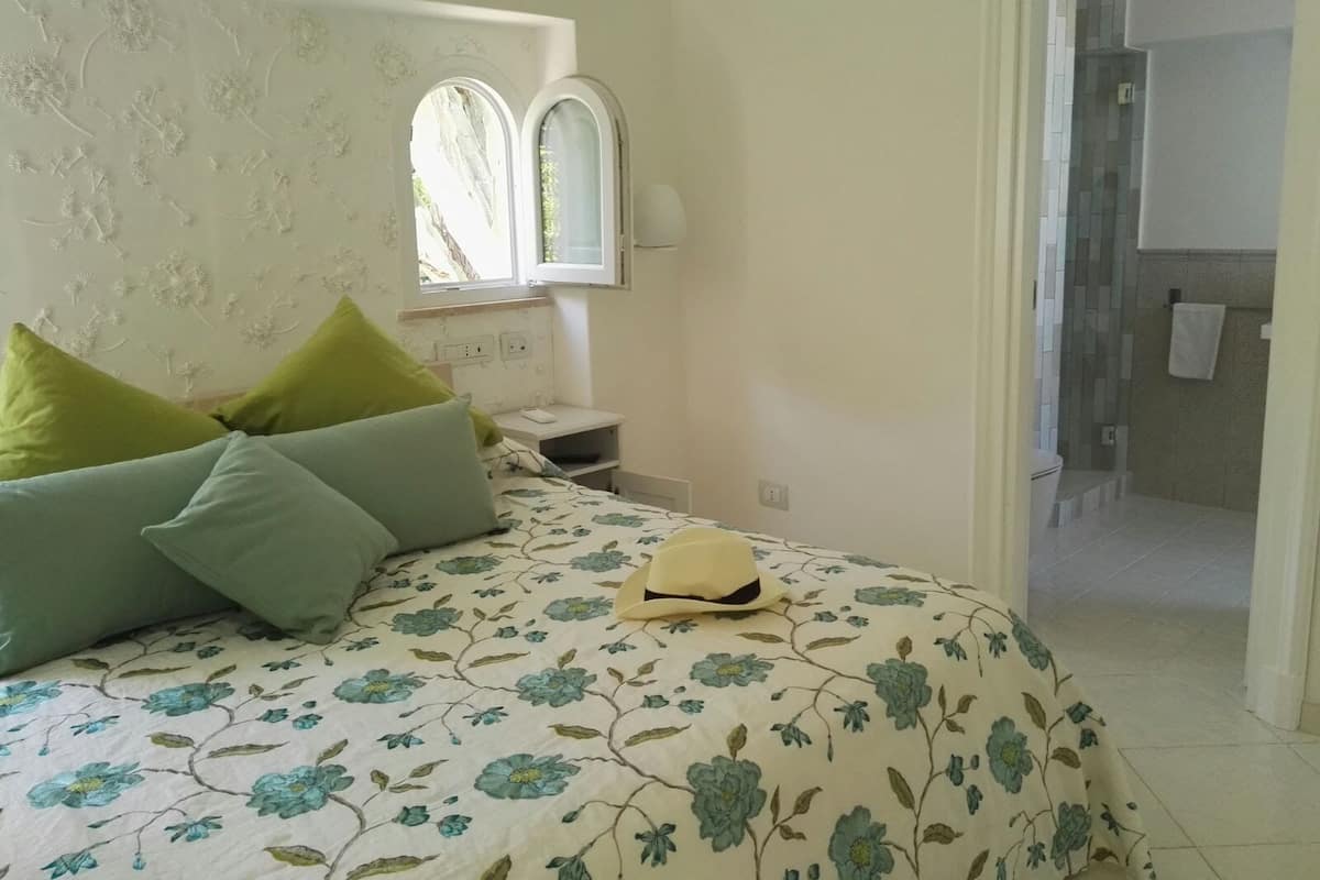 Un dormitorio con colcha estampada, almohadas verdes, una mesita de noche con lámpara y una ventana con vistas al exterior.