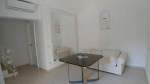 Un salón minimalista con una mesa de vidrio, un sofá blanco y una pared blanca con un ventanal pequeño.