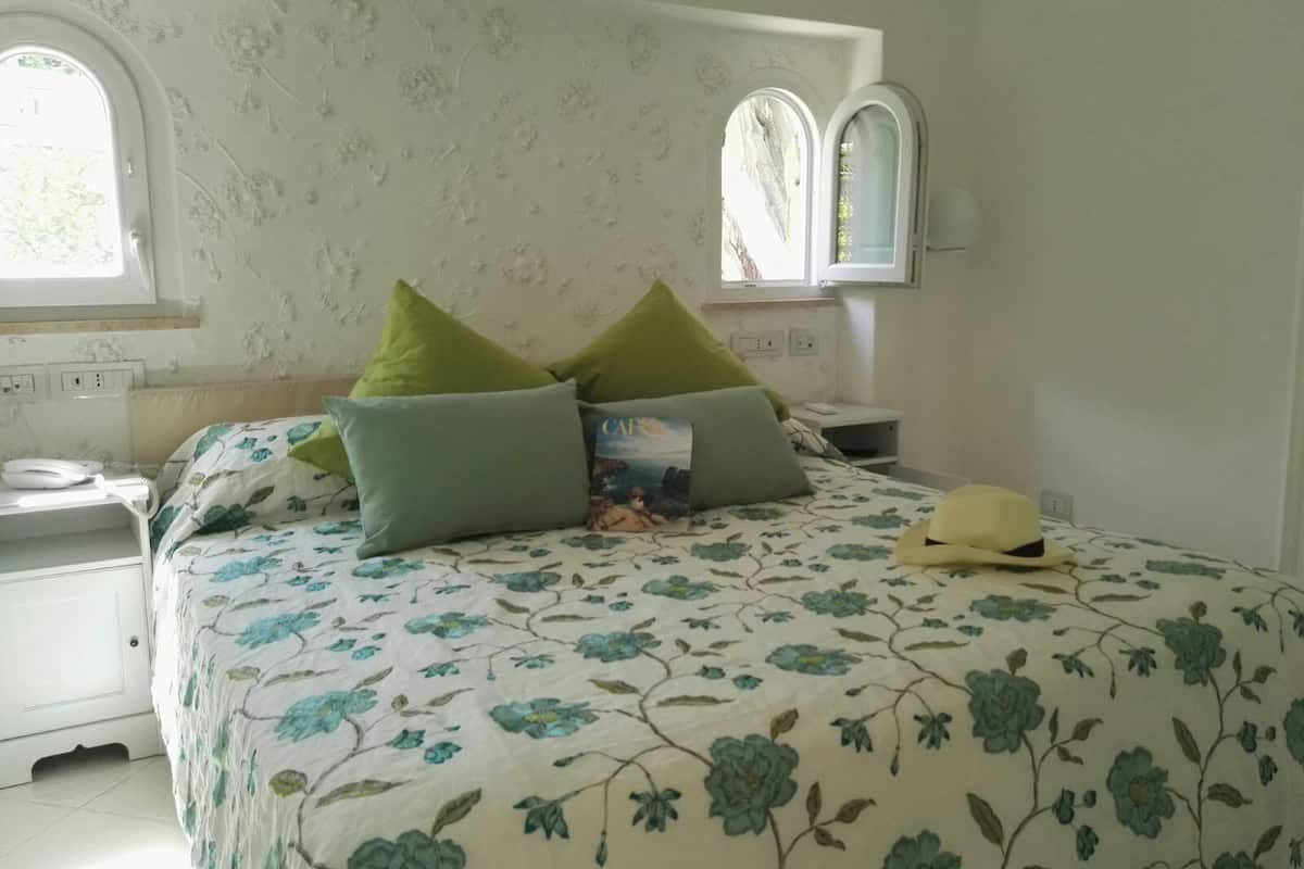 Un dormitorio con colcha estampada, dos almohadas verdes, una mesita de noche blanca, una mesilla auxiliar, un espejo y una ventana con vistas a follaje.