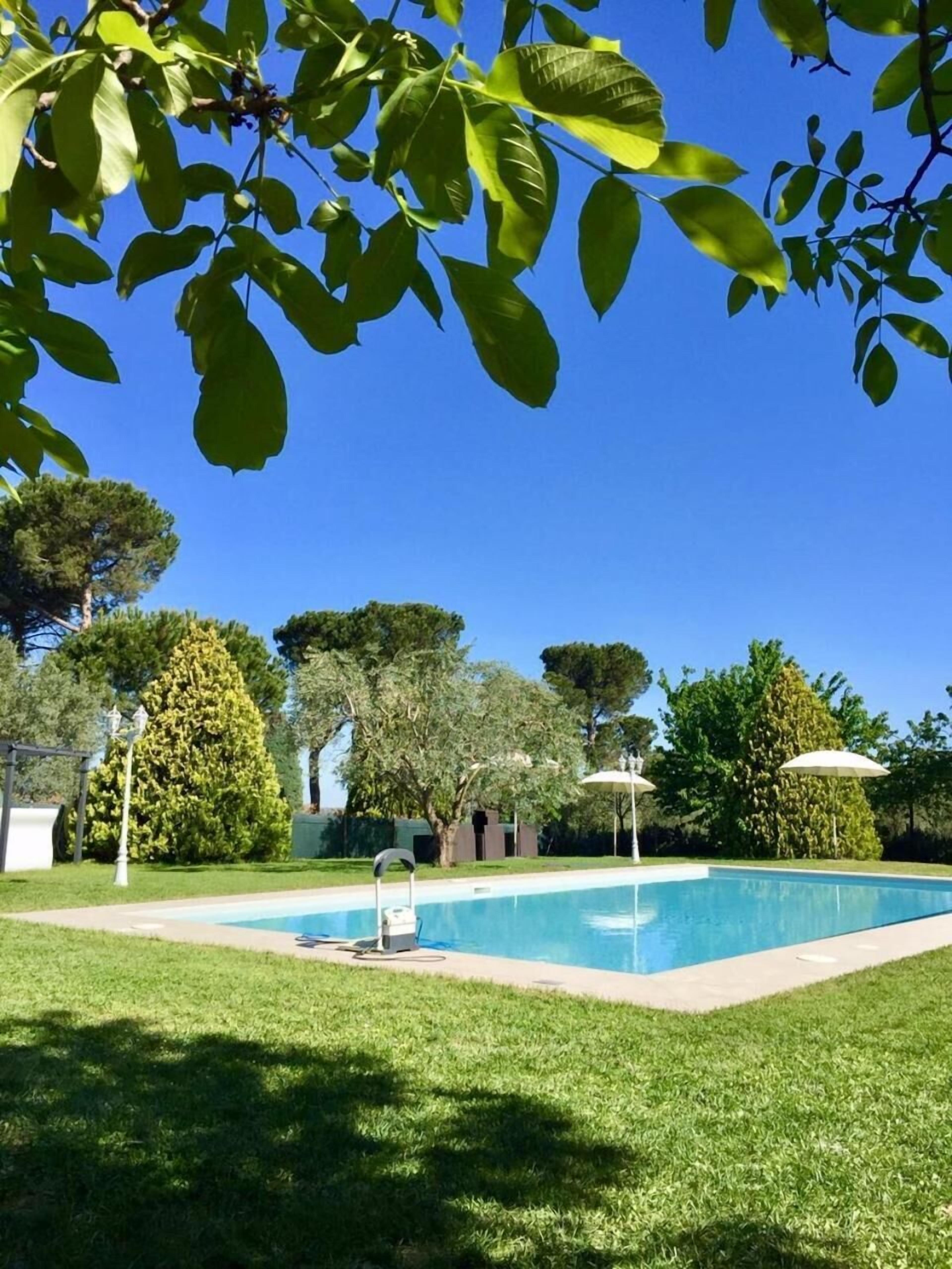 Una piscina con un cielo azul despejado, rodeada de césped y árboles bien cuidados.