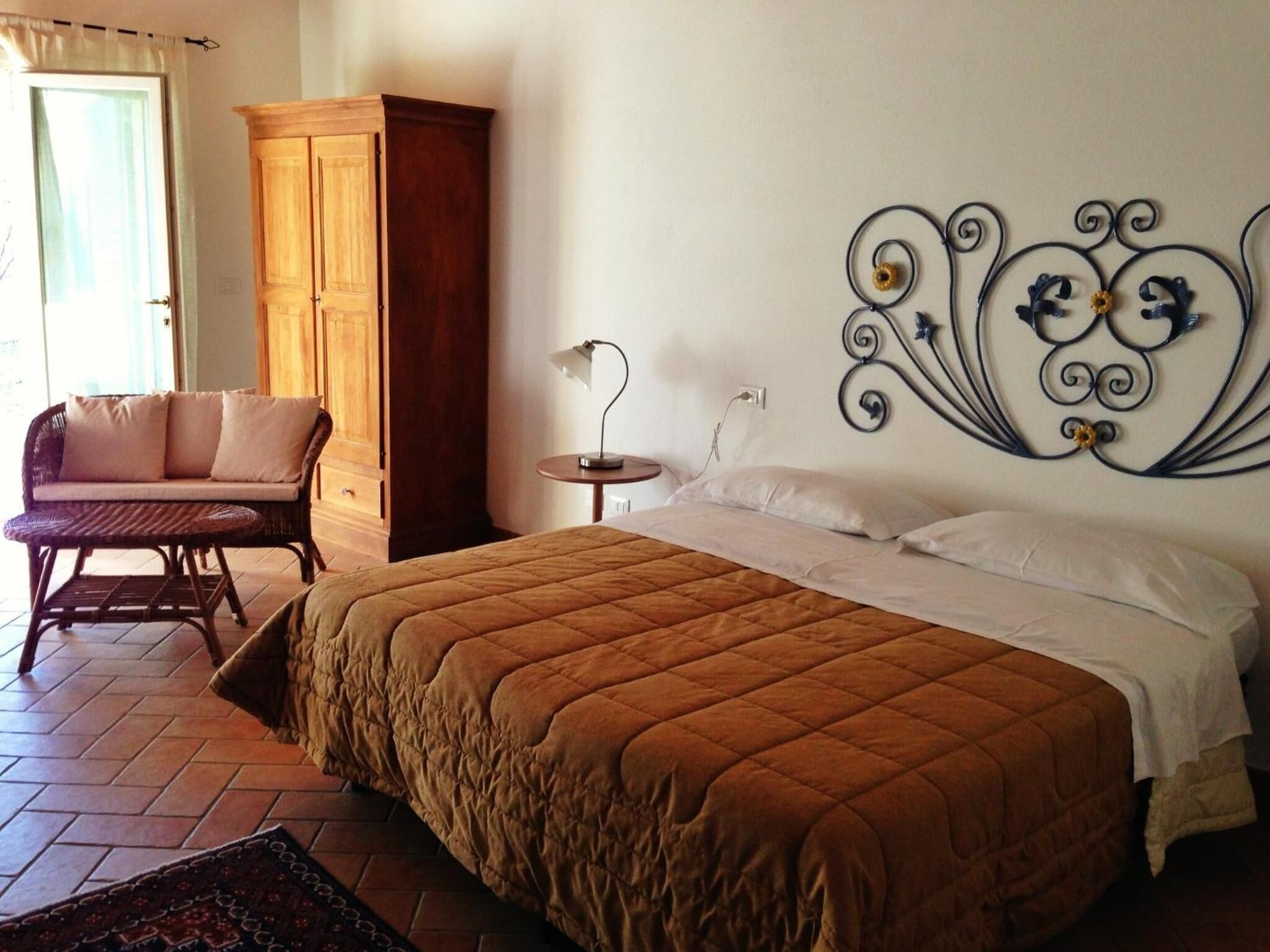 A bedroom with a large bed, a wooden wardrobe, a wicker chair, and a decorative wall piece.