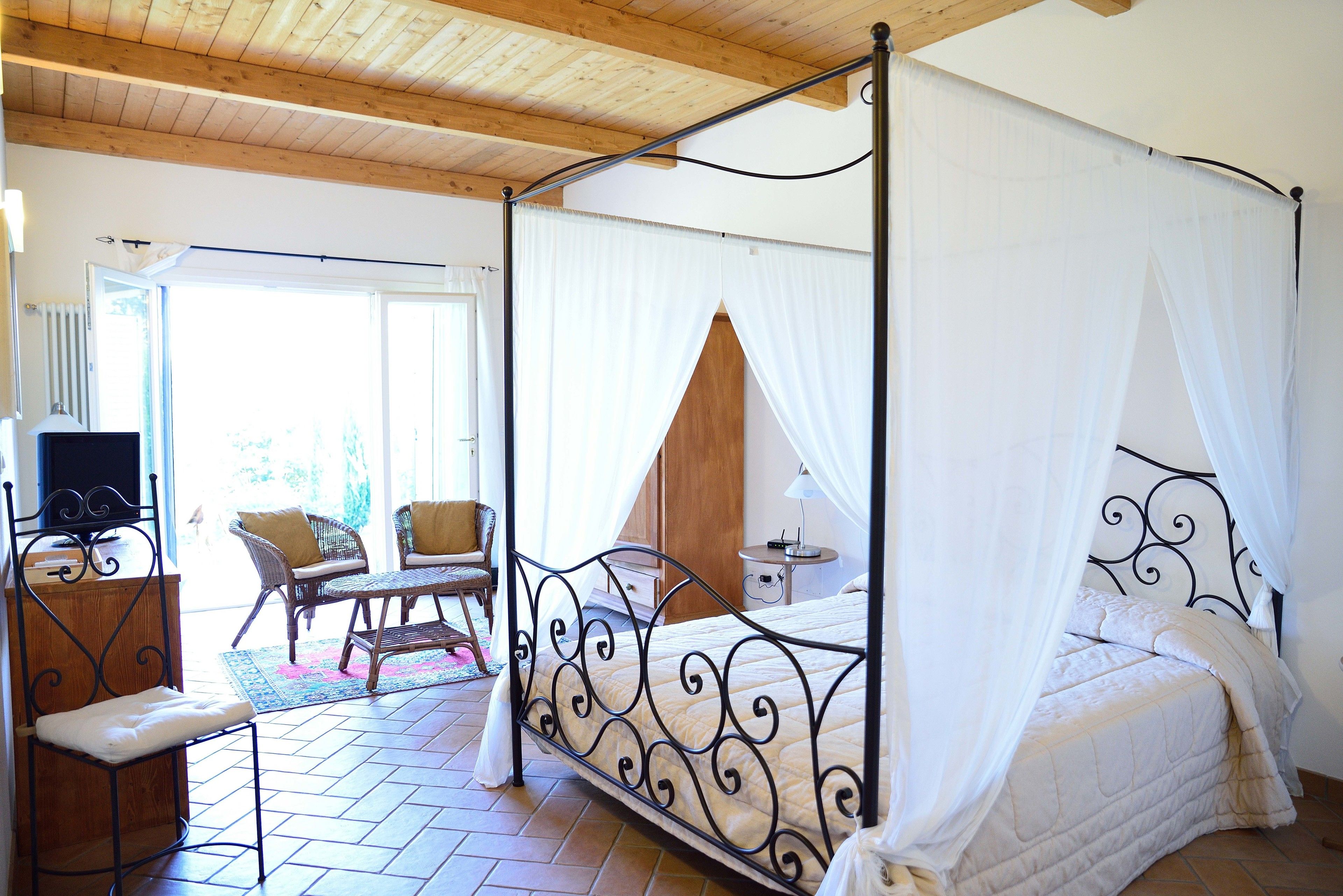 A four-poster bed with white bedding, a wooden floor, wicker furniture, and a view of the outdoors.