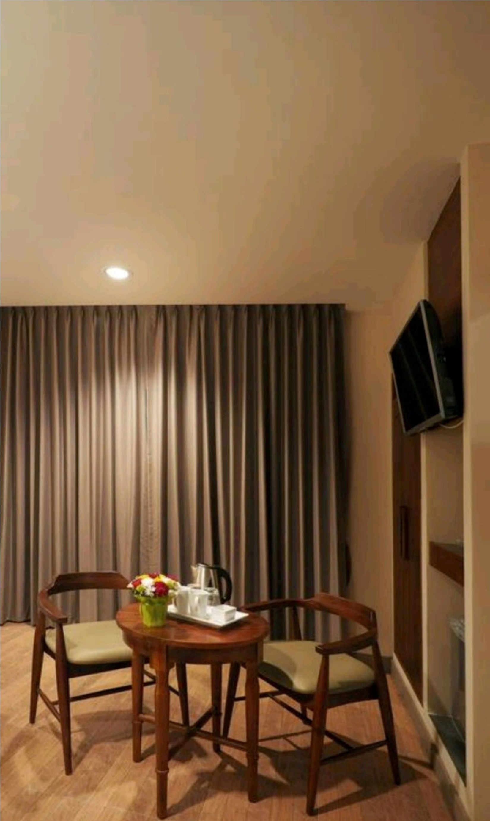 Habitación de hotel con una mesa de comedor de madera dispuesta para dos personas, un pequeño jarrón con flores y un televisor de pantalla plana montado en la pared.