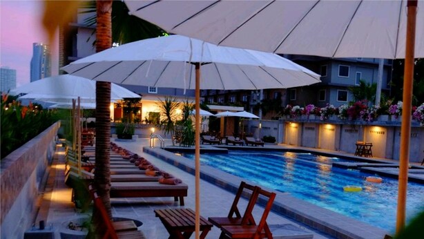 A pool area with lounge chairs and umbrellas, surrounded by buildings and plants.