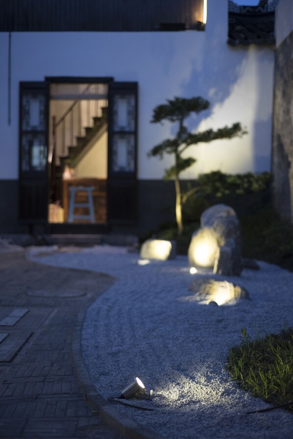 A traditional Japanese-style building with a wooden staircase leading to a room, surrounded by a gravel garden with rocks and a bonsai tree.