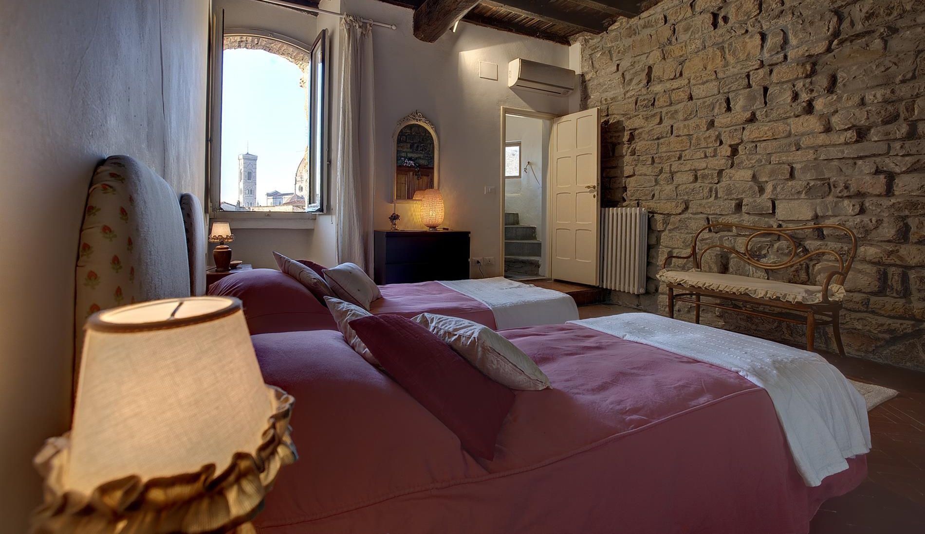 A bedroom with two beds, a stone wall, a window with a view, and a bench.