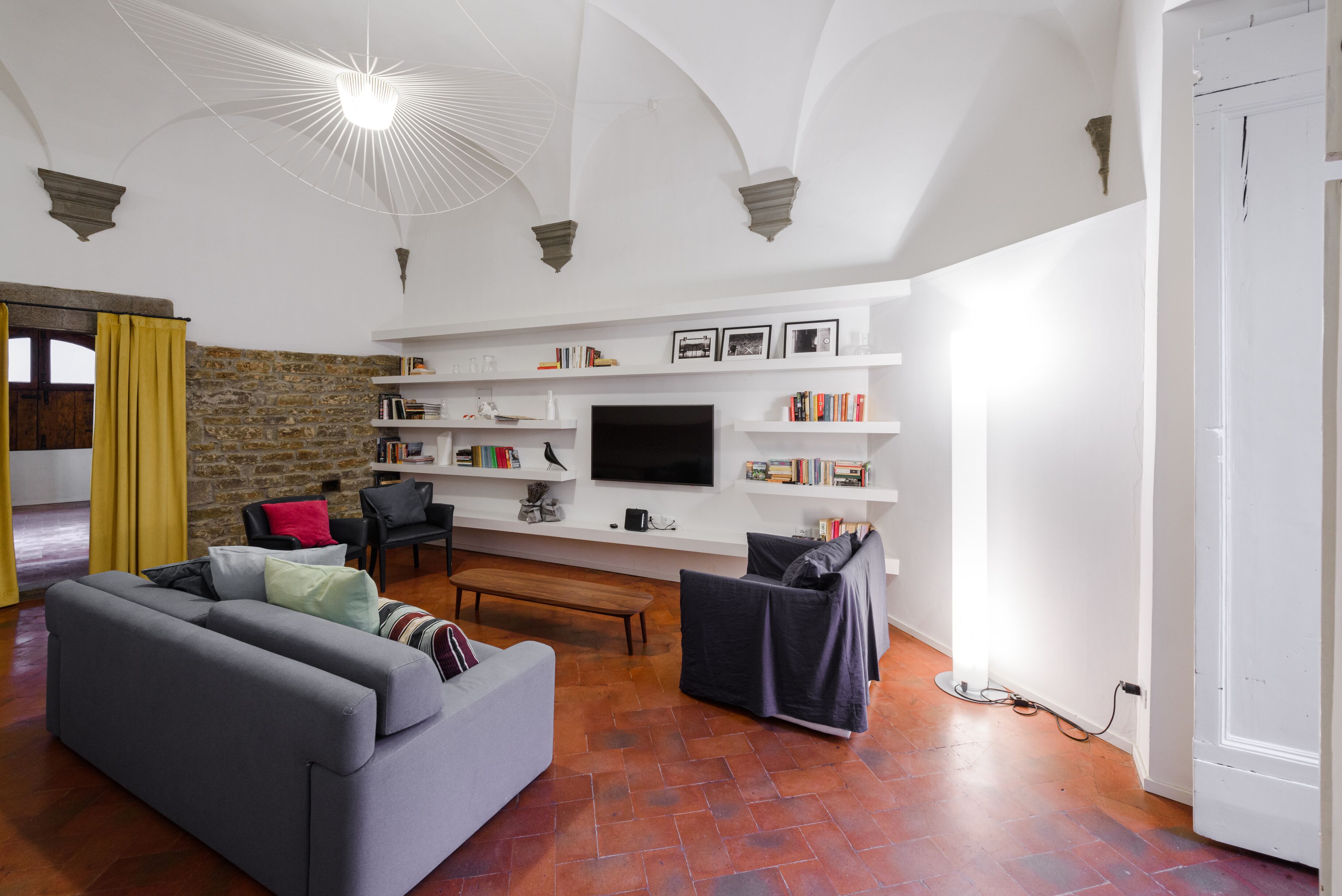 A living room with a grey sofa, a black armchair, a wooden bench, and a TV mounted on the wall. There are bookshelves with books and decorative items, and a stone wall in the background.