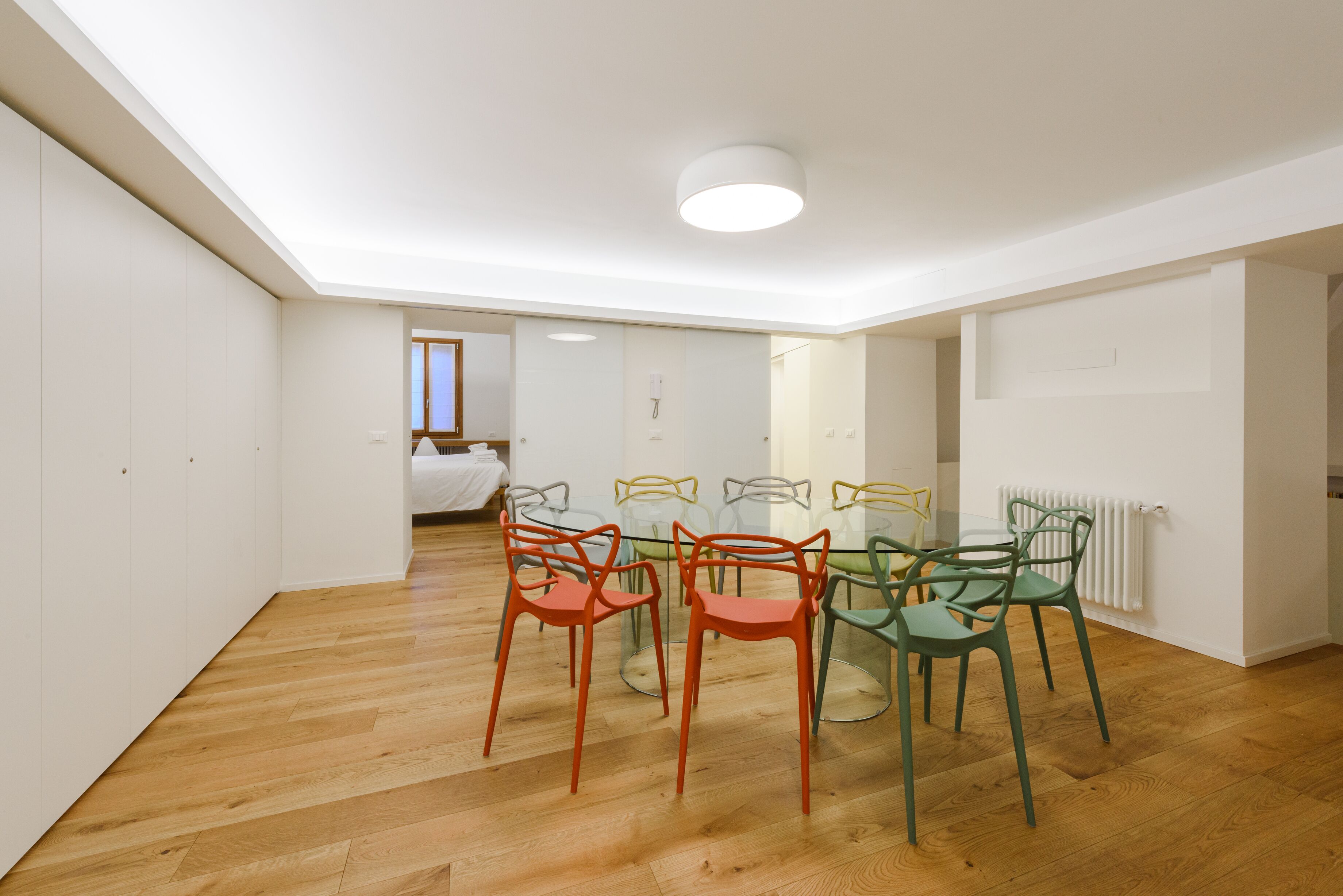 Um quarto moderno e bem iluminado, com piso de madeira, uma mesa de vidro para refeições rodeada por cadeiras coloridas, e uma cama visível ao fundo.