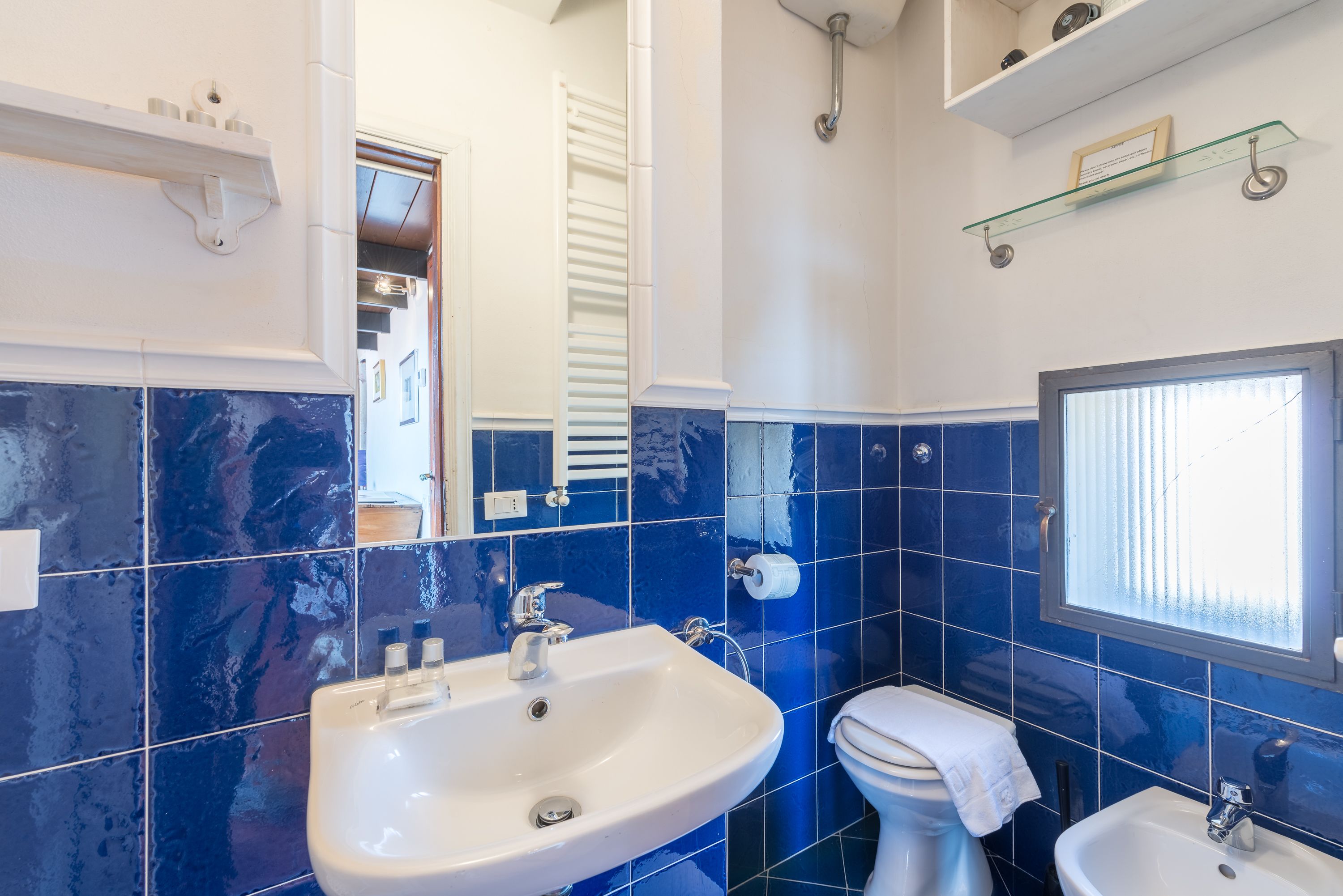 A bathroom with a white sink, a toilet, and a bidet. There is a window with blinds, a mirror, and a shelf with toiletries.