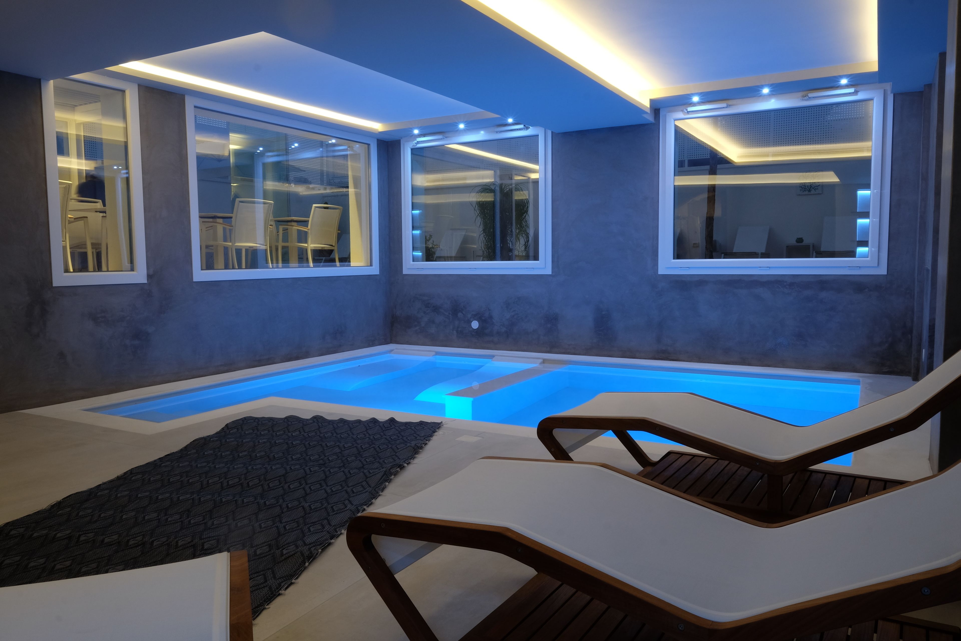 A modern indoor pool area with clear glass walls, lounge chairs, and a small seating area.