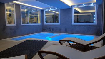 A modern indoor pool area with clear glass walls, lounge chairs, and a small seating area.