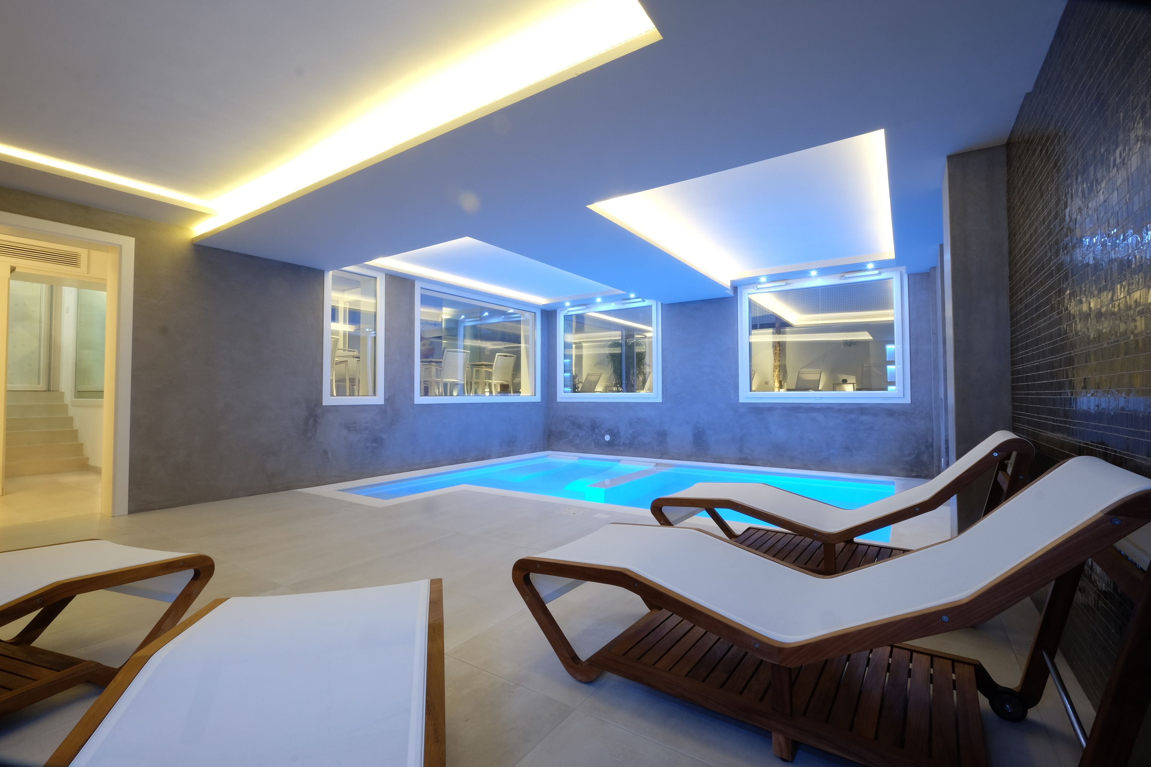 A modern indoor pool area with lounge chairs, glass walls, and recessed lighting.