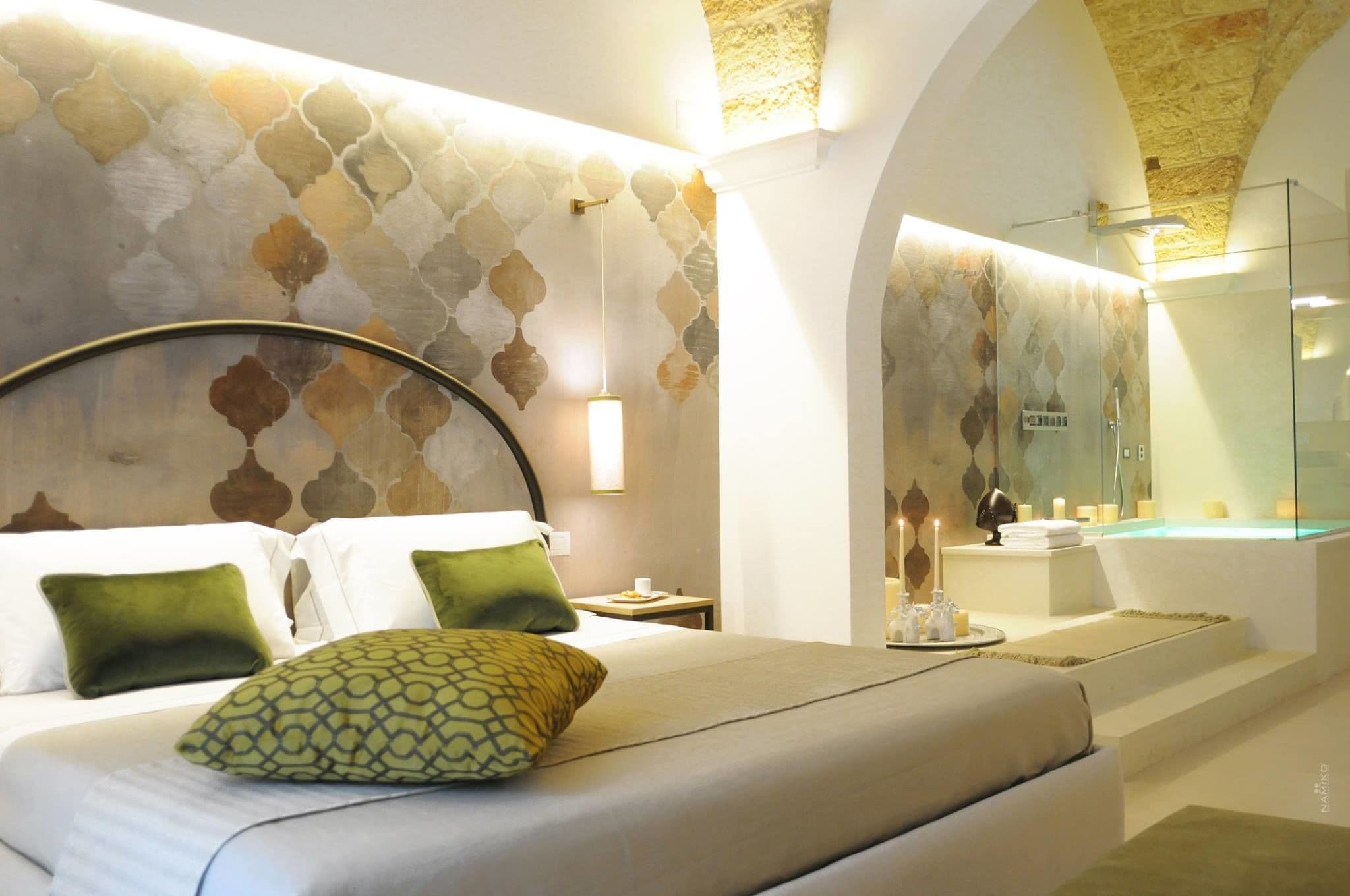 A hotel room with a bed, a nightstand, a glass-enclosed bathroom with a bathtub, and a unique patterned wall.
