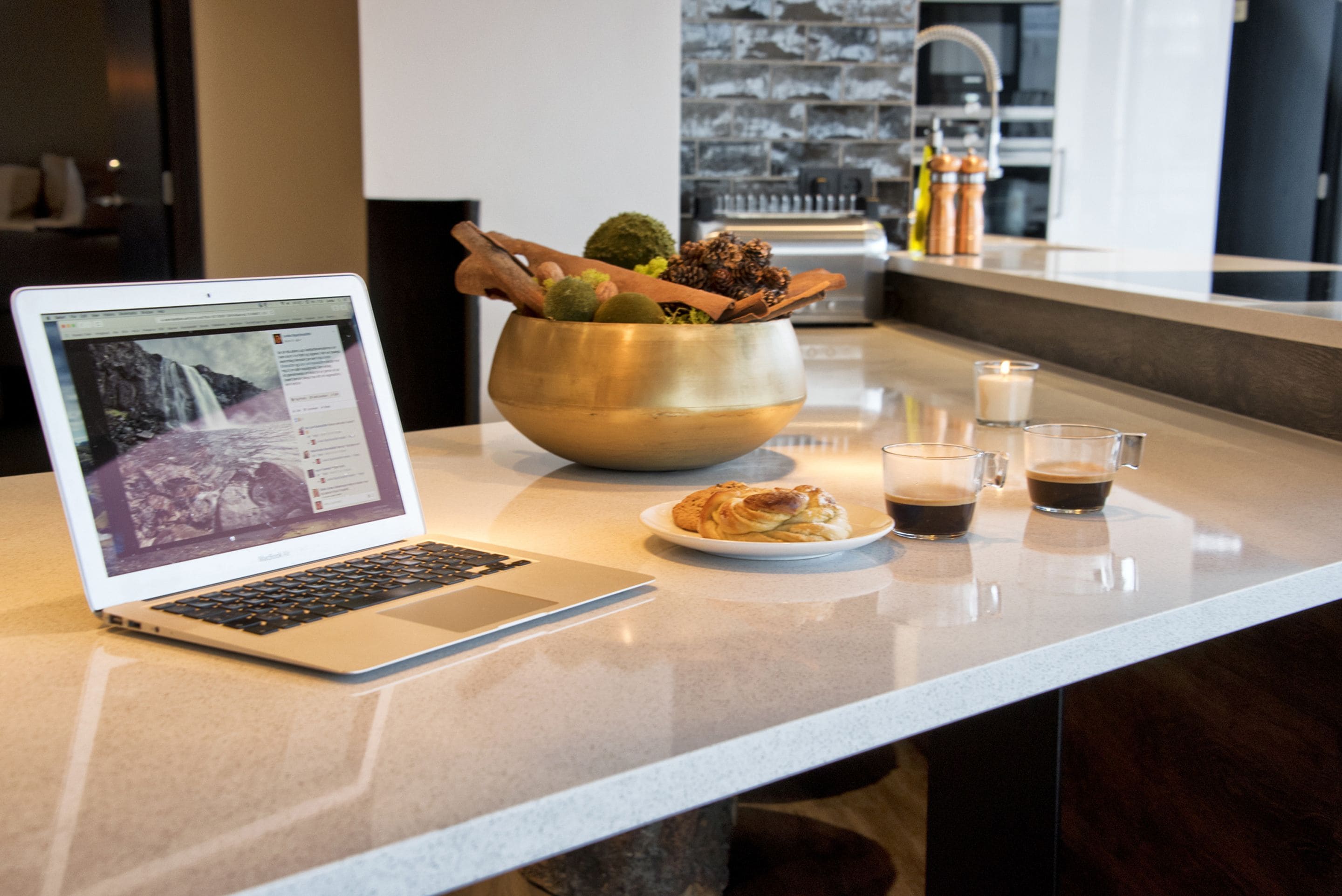 Ein Laptop steht auf einer Küchenarbeitsplatte neben einer Obstschale und einem Croissant.