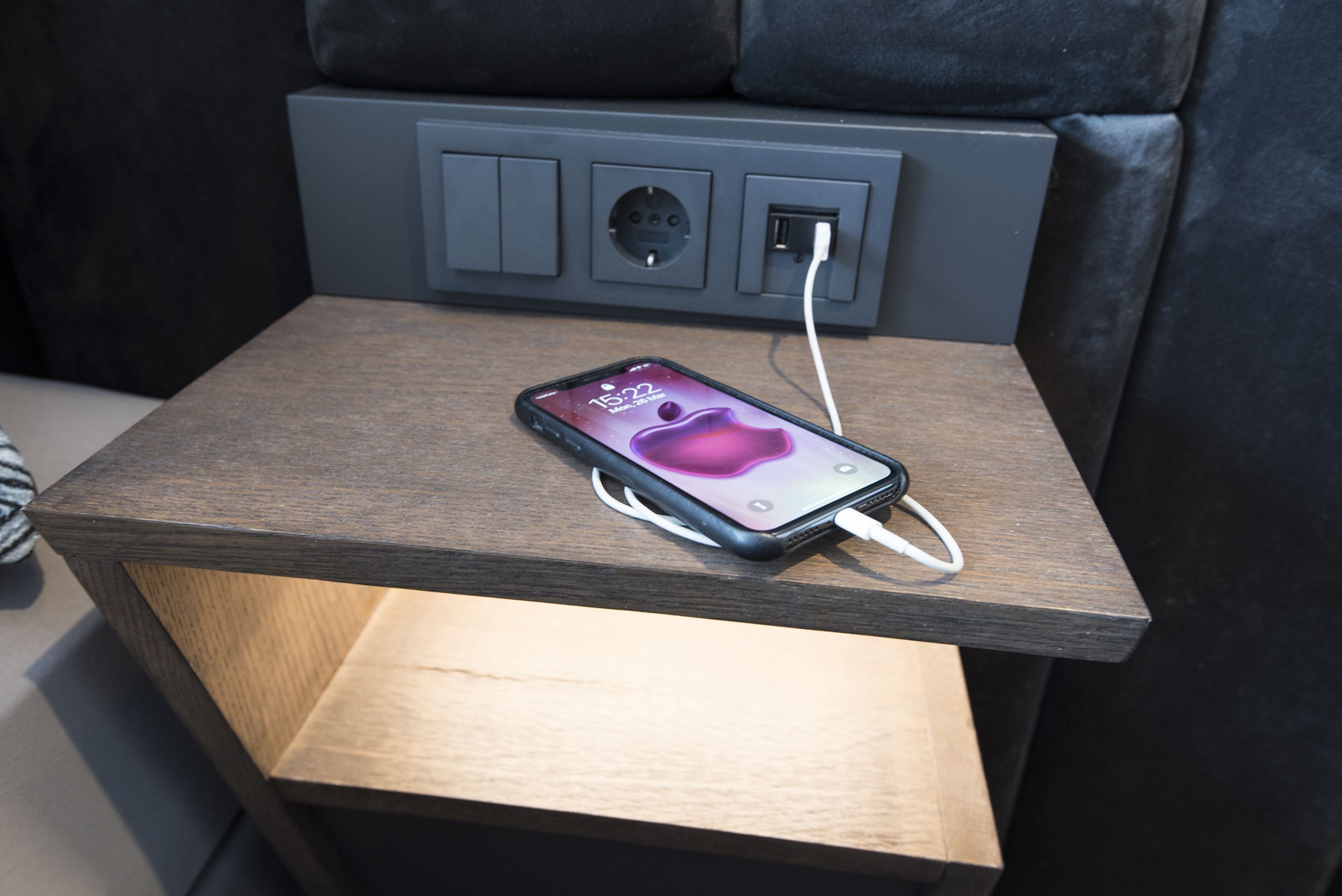 Un teléfono inteligente cargándose sobre una mesita de noche de madera, con un enchufe.