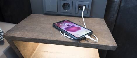 Un teléfono inteligente cargándose sobre una mesita de noche de madera, con un enchufe.