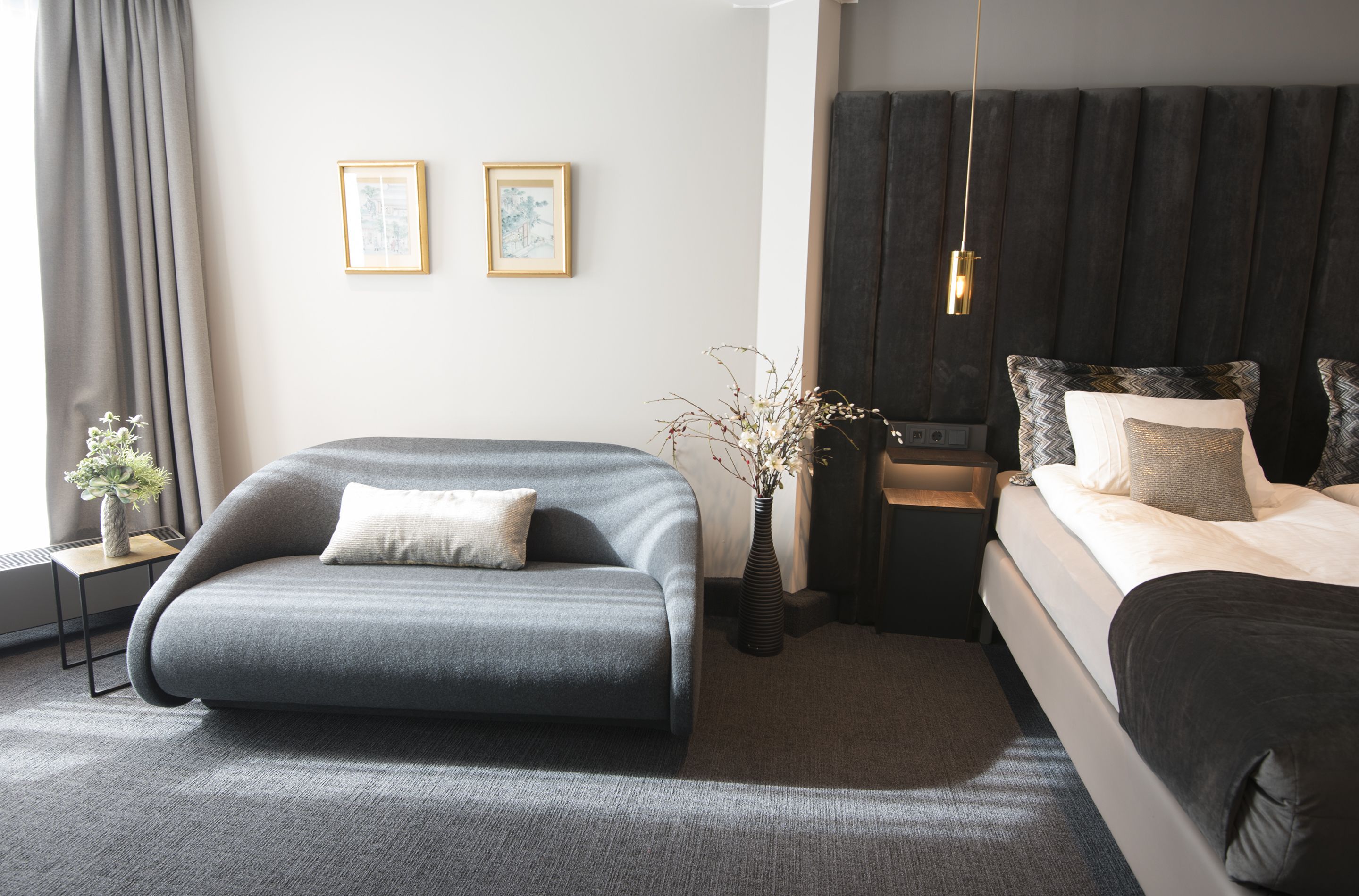 A modern hotel room with a grey sofa, a bed with patterned pillows, a nightstand, and two framed pictures on the wall.