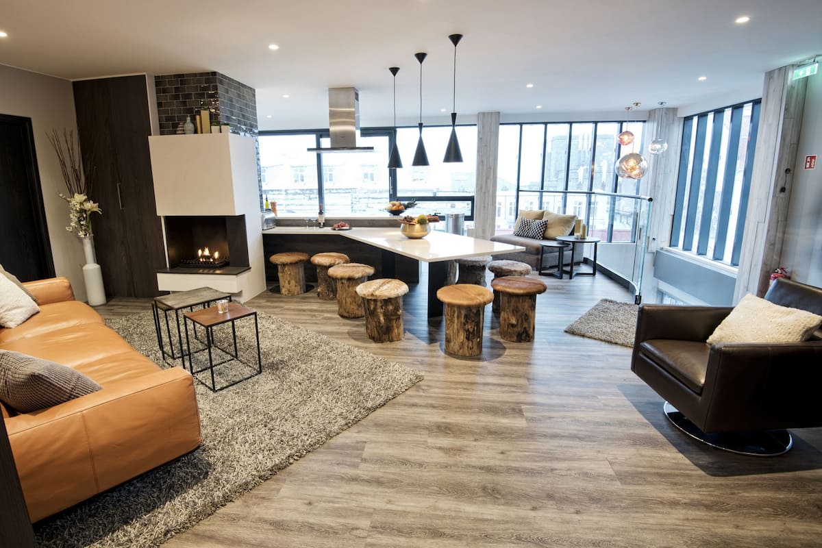 A modern living room with a fireplace, wooden stools, a sofa, and a dining area with a large window offering a city view.