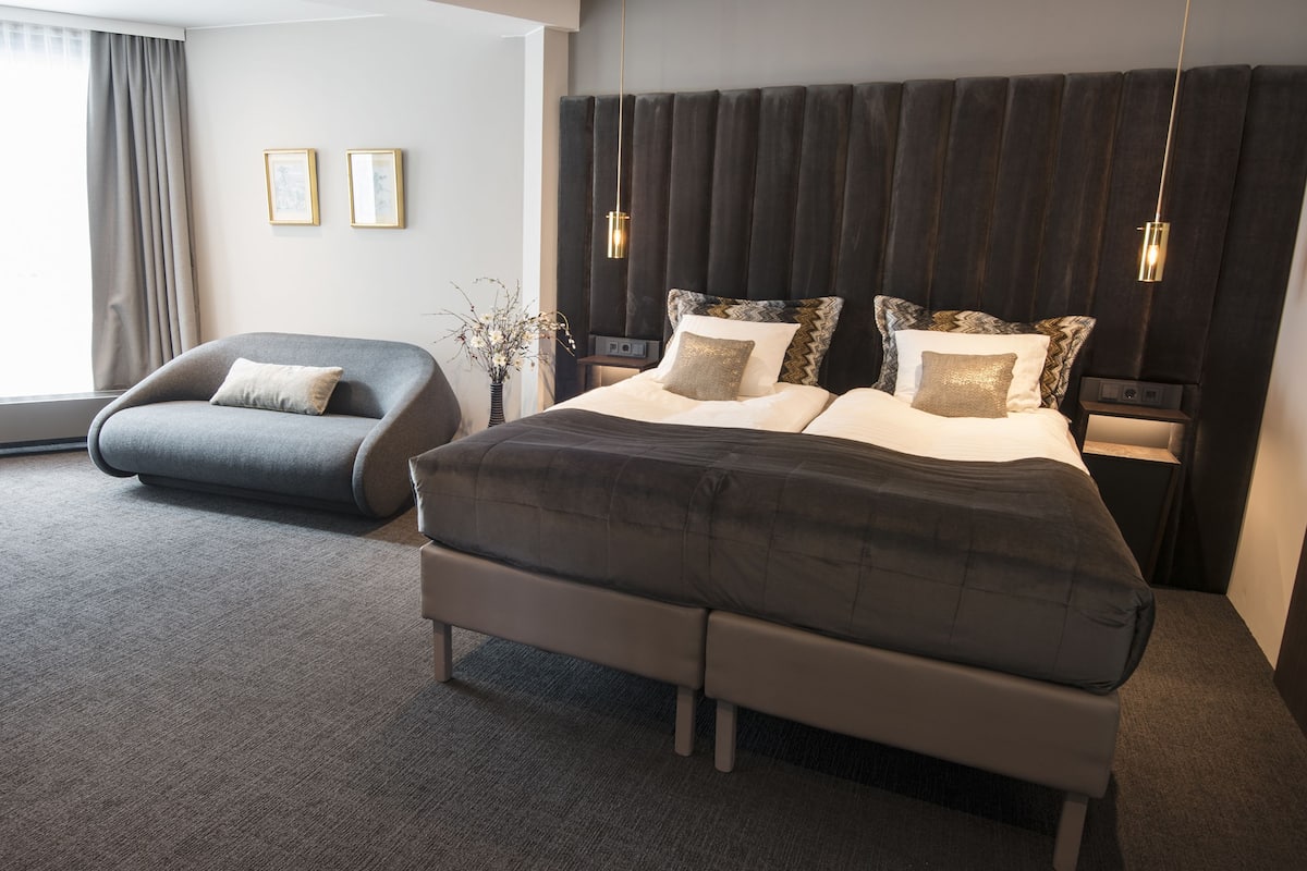 A modern bedroom with a large bed, a grey armchair, and a bedside table with a lamp.