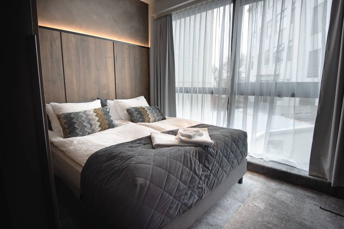 A modern hotel room with a large bed, a wooden headboard, and a view of the city through sheer curtains.