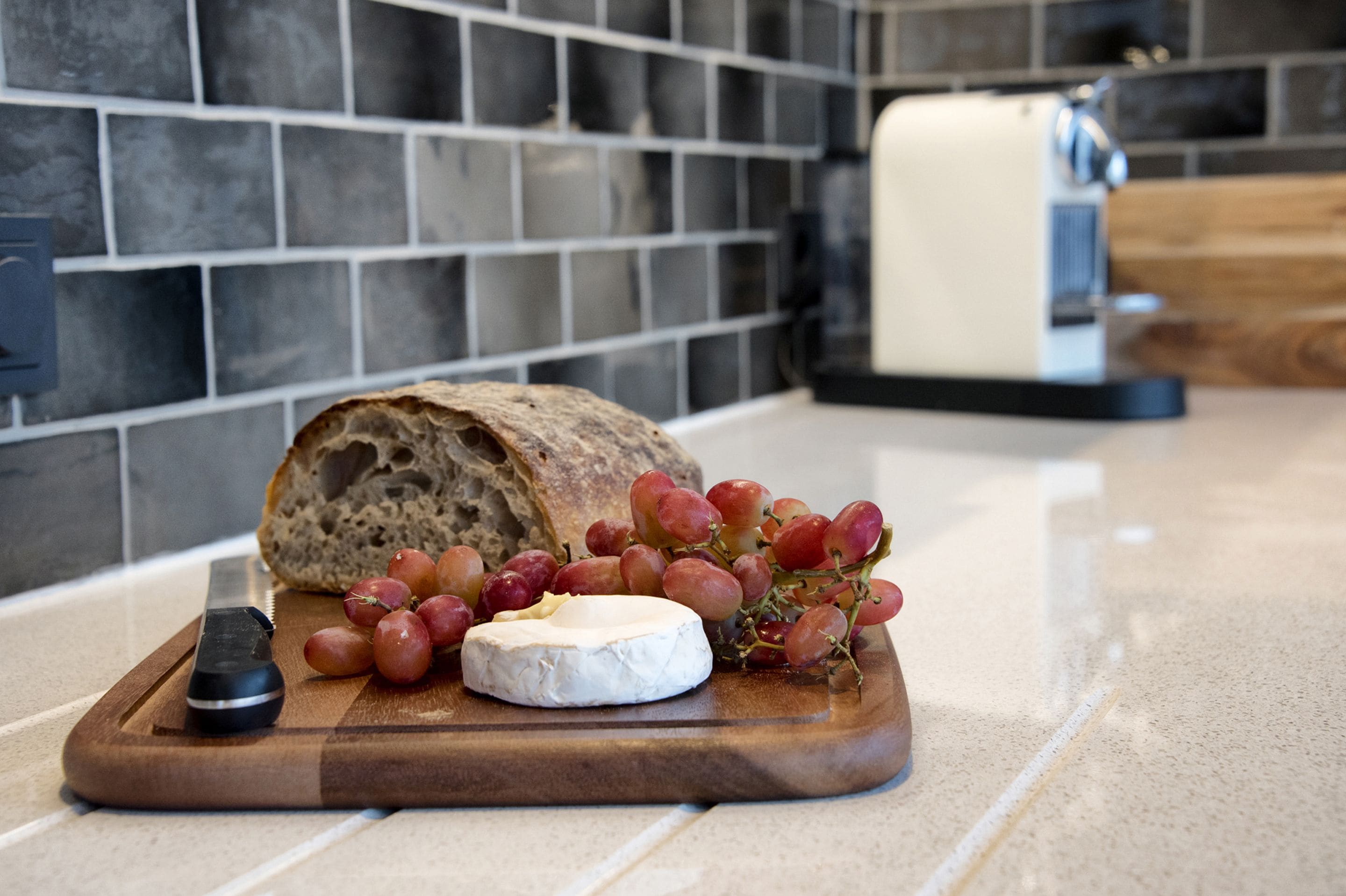 A wooden cutting board with a loaf of bread, a piece of cheese, and a bunch of grapes on a kitchen counter.