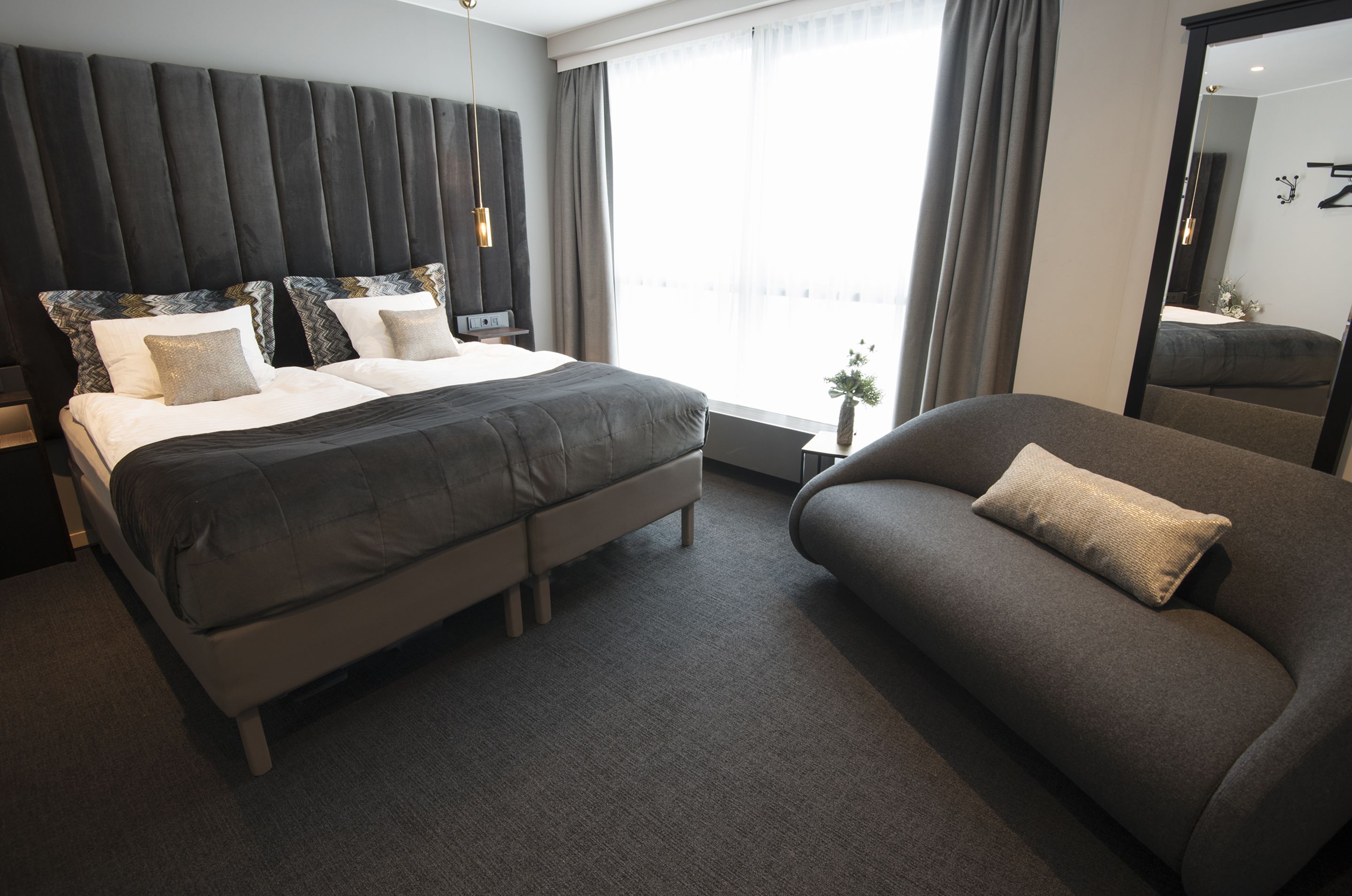 Quarto de hotel moderno com uma cama grande, um sofá e uma janela ampla com cortinas.