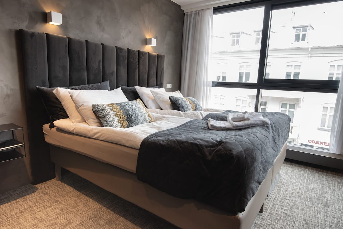 A modern hotel room with a large bed, a nightstand, a window with a view of buildings, and a wall-mounted light fixture.