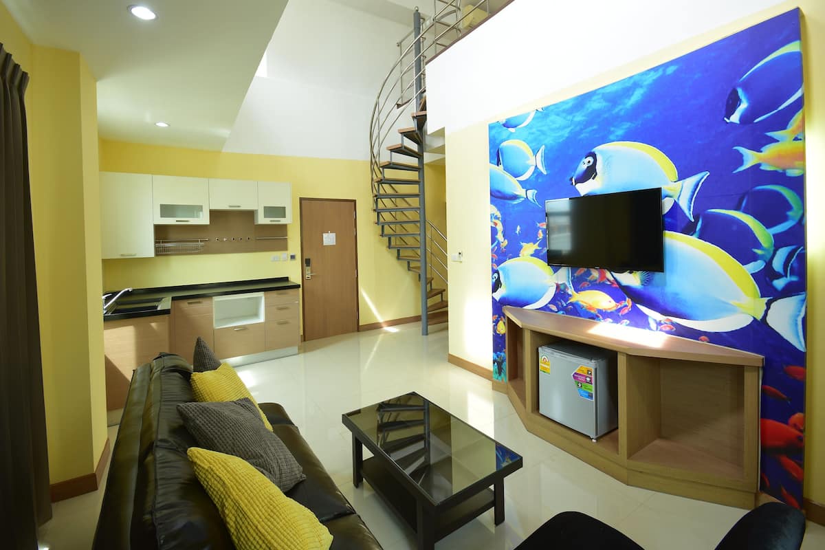 Een moderne woonkamer met een bank, salontafel en een aan de muur bevestigde televisie. Achtergrond toont een keuken met een trap naar een hoger niveau.