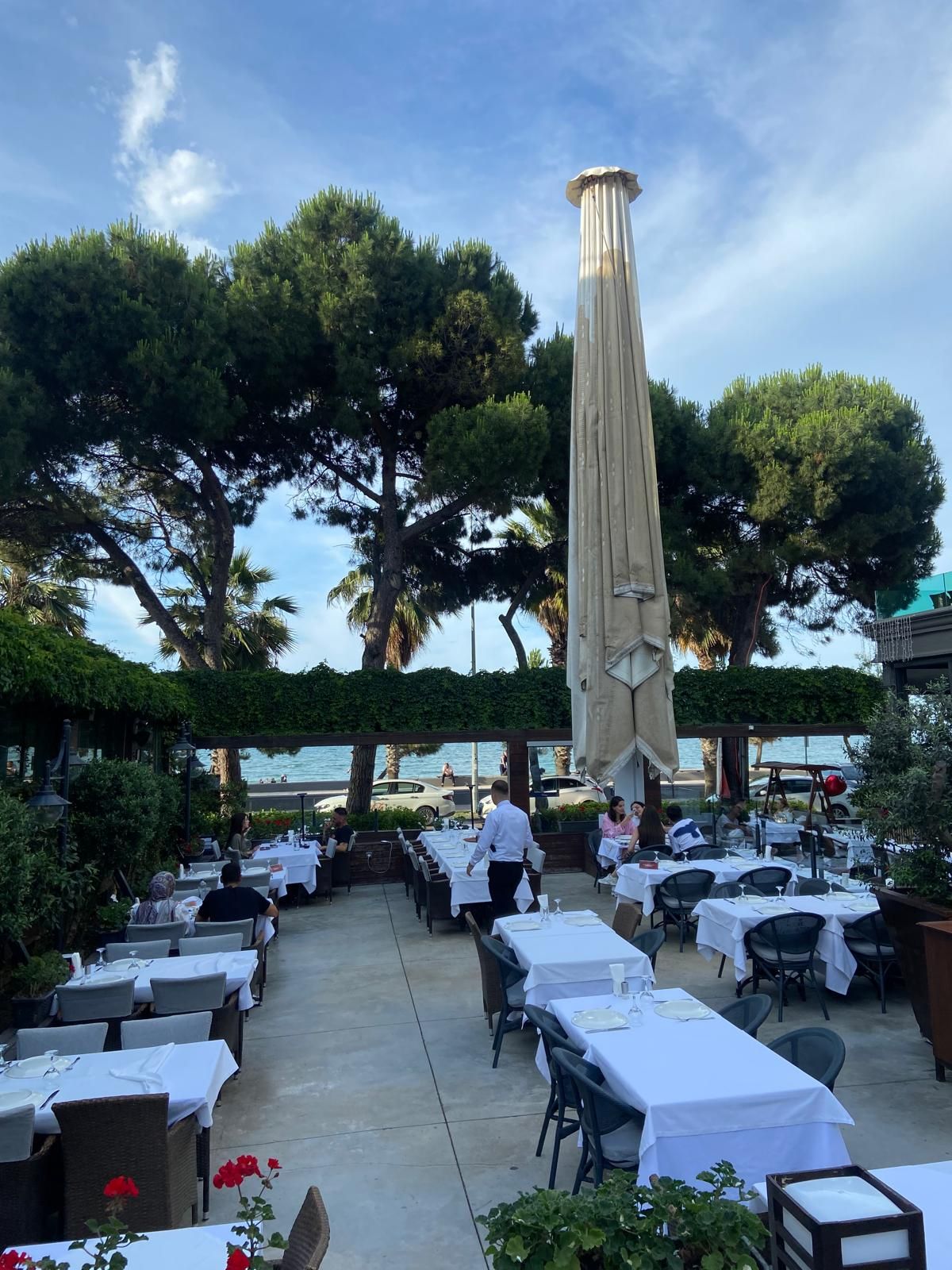 An outdoor dining area with tables and chairs, a tall column, and a view of the sea.