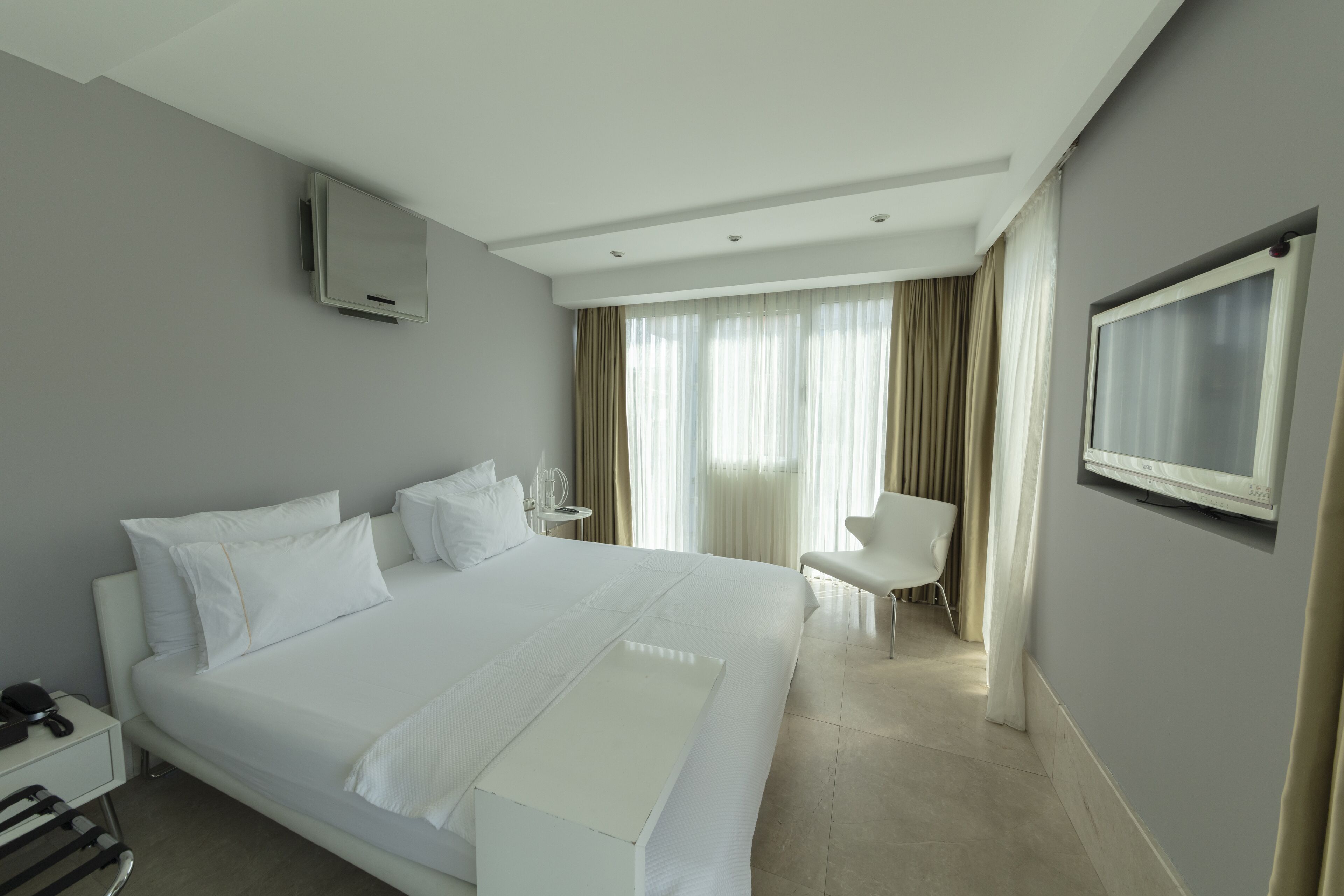 A modern hotel room with a large bed, a bedside table, a chair, a TV mounted on the wall, and a window with curtains.