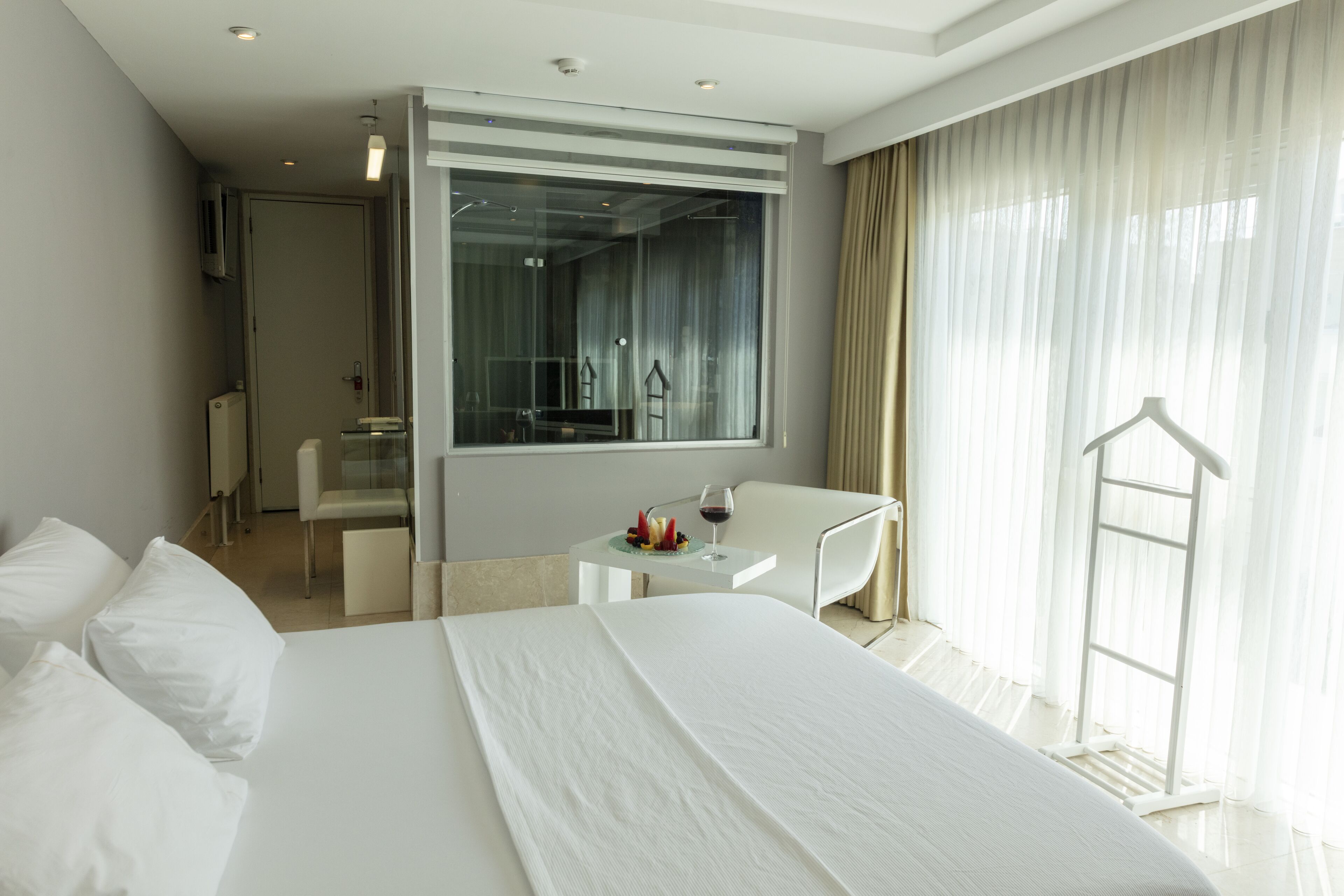 A modern hotel room with a large bed, a small desk with a chair, a mirror, and a view of the outdoors.
