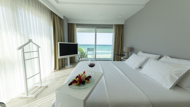 Habitación de hotel con una cama grande, televisión, una mesa de comedor con fruta y vino, y vista al mar por la ventana.